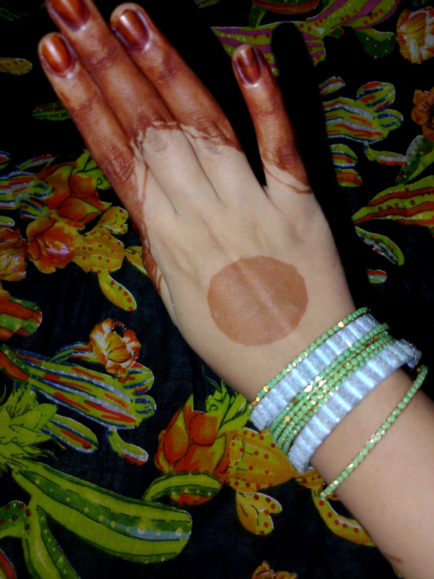 The image shows a close-up of a person's hand adorned with henna and bracelets, resting on a dark fabric with a colorful floral and vegetable print. The hand has dark brown henna applied to the fingertips and a circular design on the palm. Several bracelets, including beaded ones with green and white elements and gold accents, are worn on the wrist. The fabric underneath features a repeating pattern of stylized yellow flowers with red dots, and green elongated shapes resembling cucumbers or gourds, against a black background. The overall impression is of a decorative or celebratory adornment. There is no identifiable location in the city of Thane, India, depicted in the image.