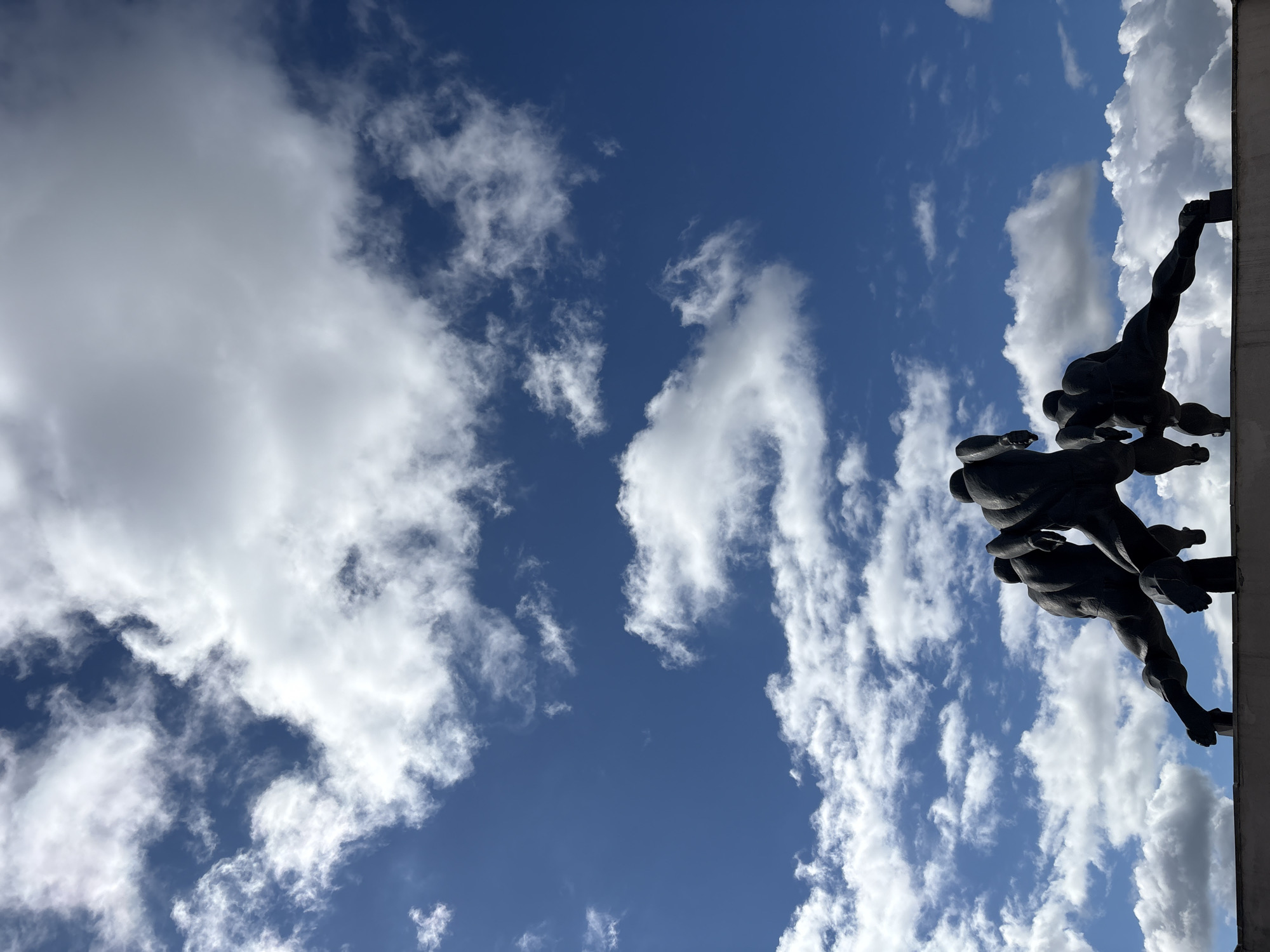 The image depicts a low-angle view of a cloudy blue sky with a portion of a dark-colored monument in the lower-right corner. The monument shows figures in a dynamic pose, possibly laborers or athletes, suggesting strength and unity. The style of the monument, combined with the location's architecture, suggests it is located in Minsk, Belarus. The scene is captured under daylight conditions, as indicated by the well-lit sky and the shadows on the monument.