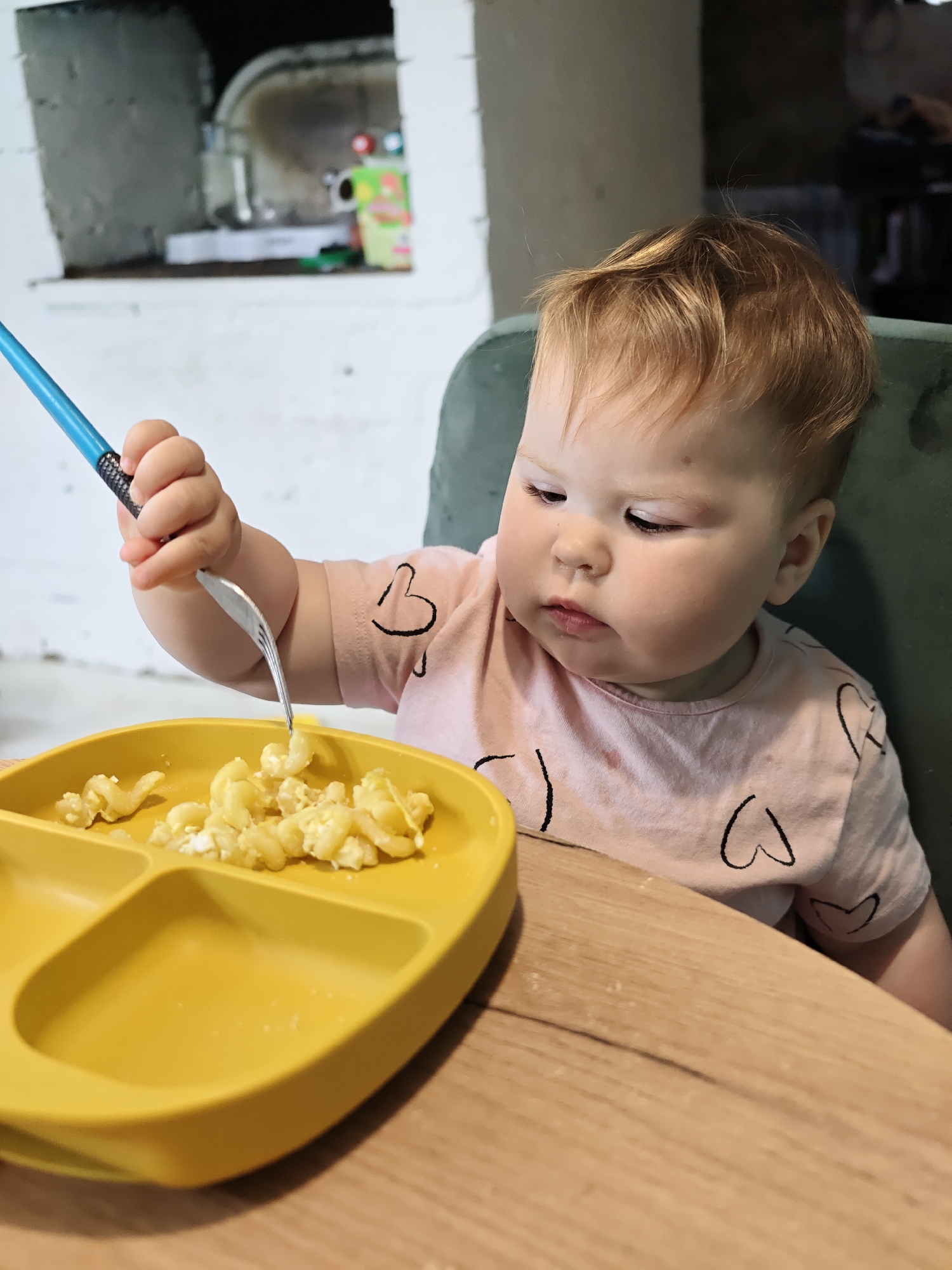 A baby sits at a wooden table in an interior space, eating macaroni and cheese off of a yellow segmented plate using a fork with a blue handle. The baby has light skin, reddish-blonde hair, and wears a pink t-shirt with black heart outlines. Behind the baby, a green chair is visible and further back, a stone structure with a dark opening can be seen. There is no location information specifying Noviy, Russia.