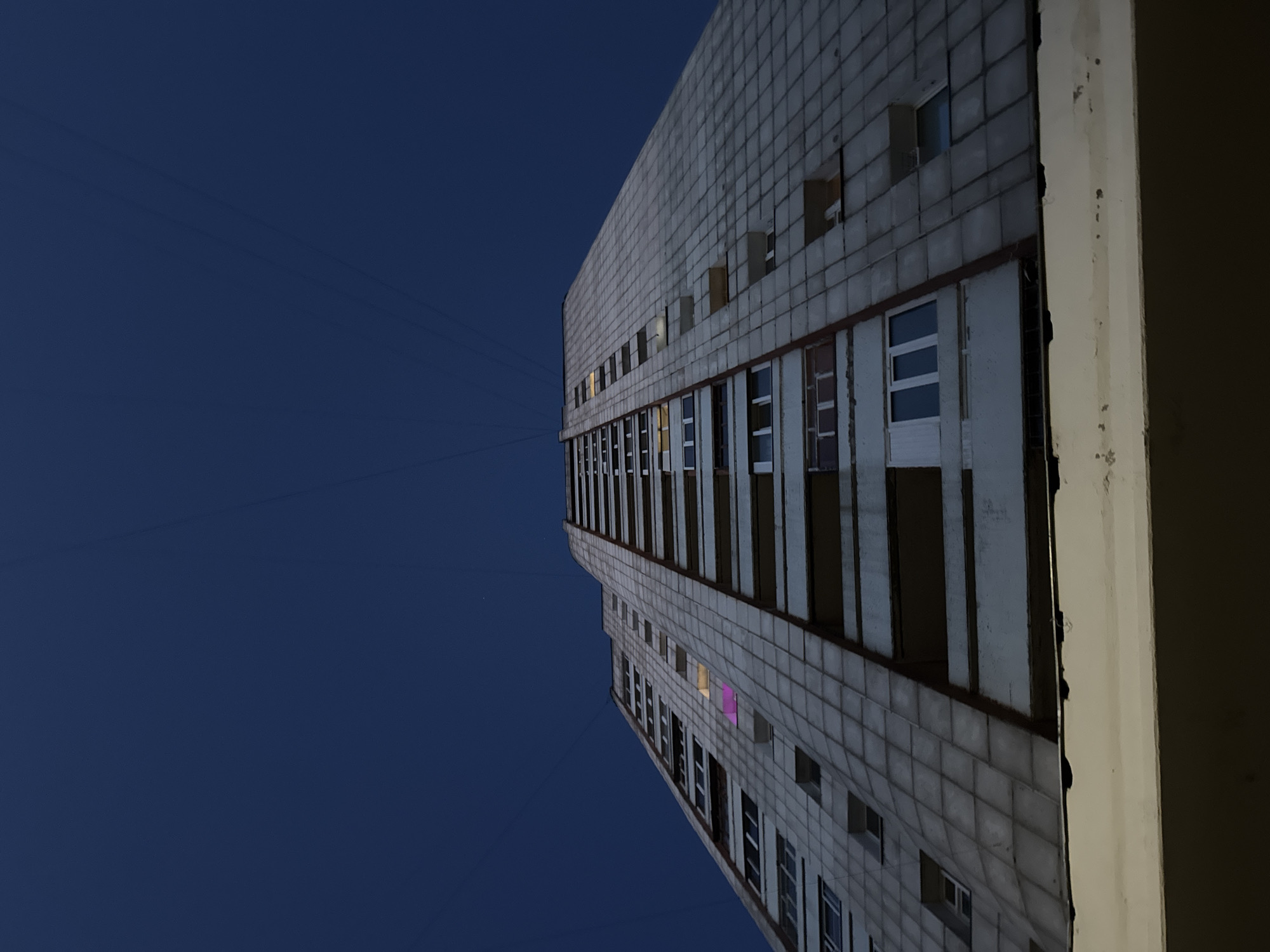 The image displays a tall, utilitarian building against a dark blue night sky. The perspective is from a low angle, looking upwards at the building which is tilted, extending from the bottom center towards the upper left. The building's facade is characterized by a grid pattern of light grey square tiles. Numerous rectangular windows are visible across its many floors. Several windows are illuminated from within, with some casting yellow and one emitting a distinct pink light, suggesting internal activity. Faint, thin lines, possibly electrical wires or cables, are visible against the dark sky on the left. On the far right edge of the frame, a segment of a lighter-colored, possibly stucco or concrete, wall or column is partially visible, indicating an adjacent structure or part of the building's exterior. No people are visible, and no specific event is depicted other than the quiet presence of the building at night. The time of day is clearly night or very late twilight due to the dark sky and artificial indoor lighting. There is no visible text. The location is Chaikovsky, Russia.