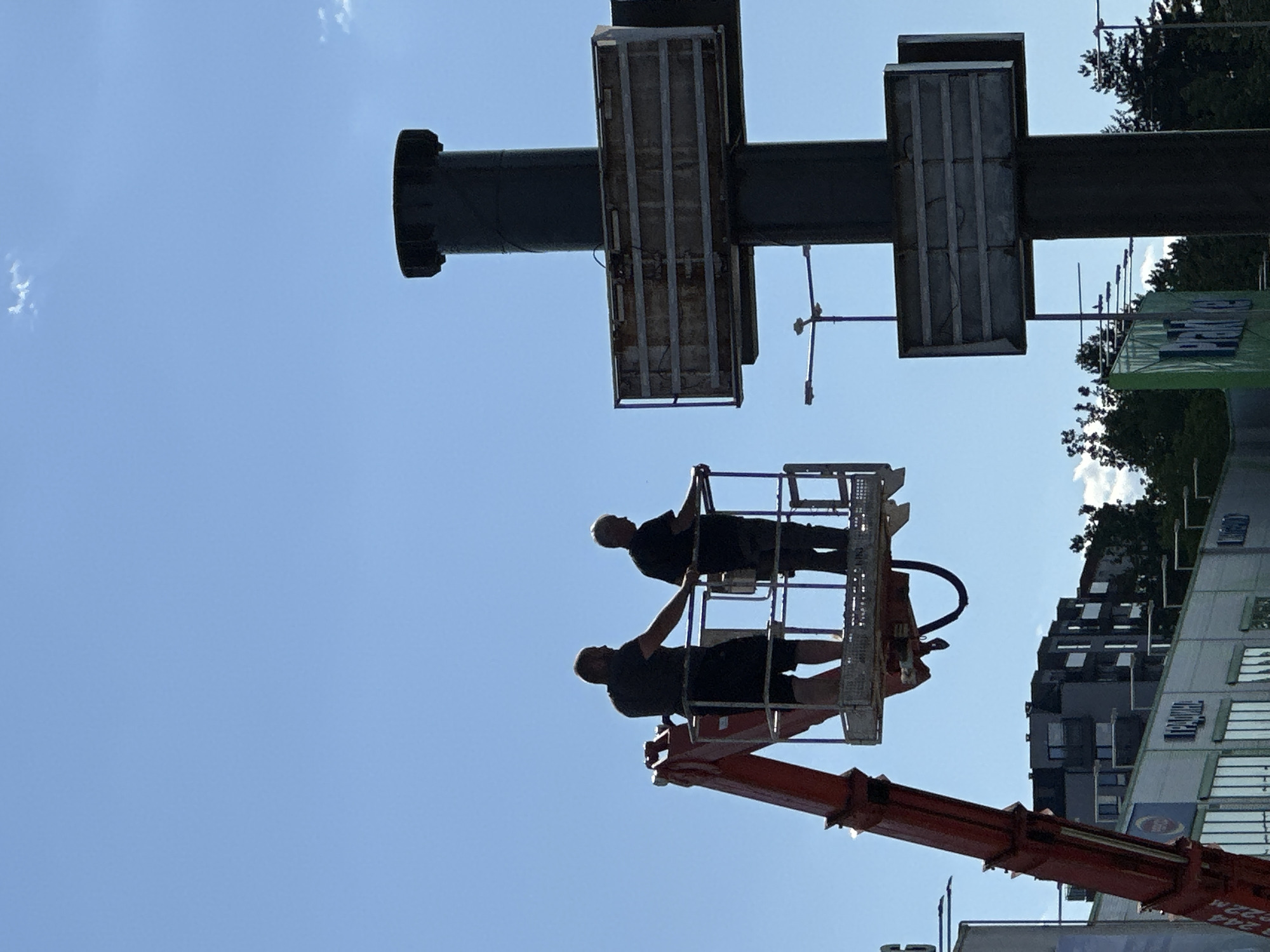 Two individuals, silhouetted against a clear blue sky, are positioned in the basket of a red aerial work platform. They appear to be engaged in work on or around a tall, dark pole that supports multiple rectangular structures, possibly signs. The pole is positioned to the right of the work platform. In the background, a modern multi-story building with dark facades and white-trimmed windows is visible, along with trees and streetlights. The overall scene suggests an outdoor urban or commercial environment during daylight. This image was taken in Veliko Tarnovo, Bulgaria.
