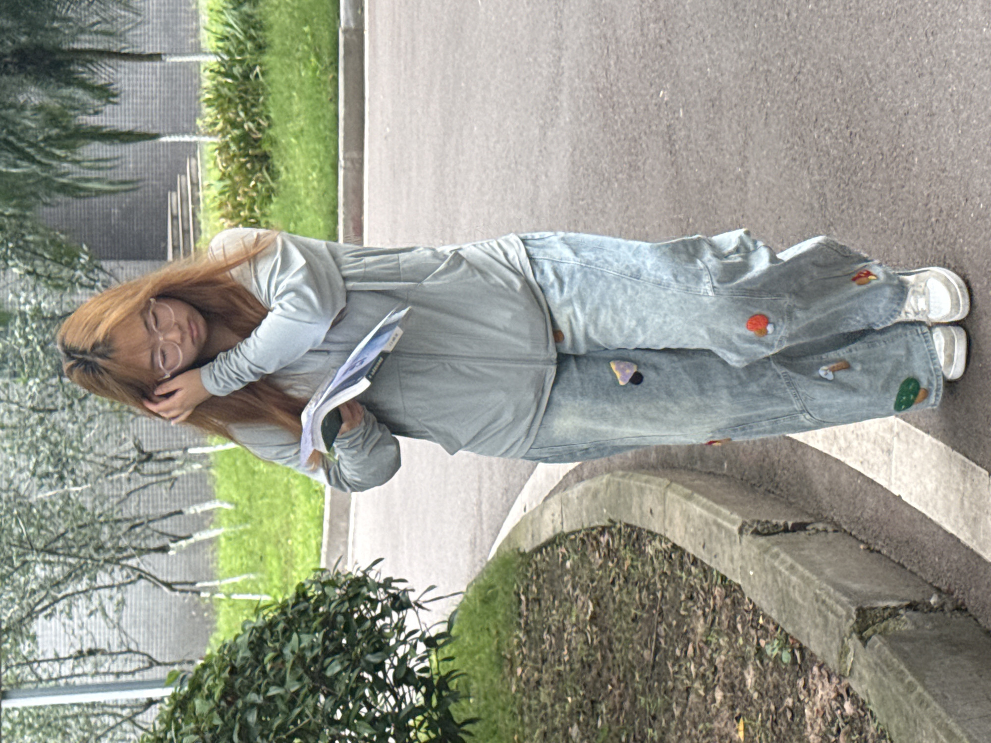 A young woman lies on a sidewalk in Jiangjin, China. She is wearing glasses, a light blue jacket, and decorated jeans, and she has light brown hair. She appears to be sleeping, with her head resting on her hand. A book or notebook is in her other hand. The sidewalk is bordered by greenery and a road.