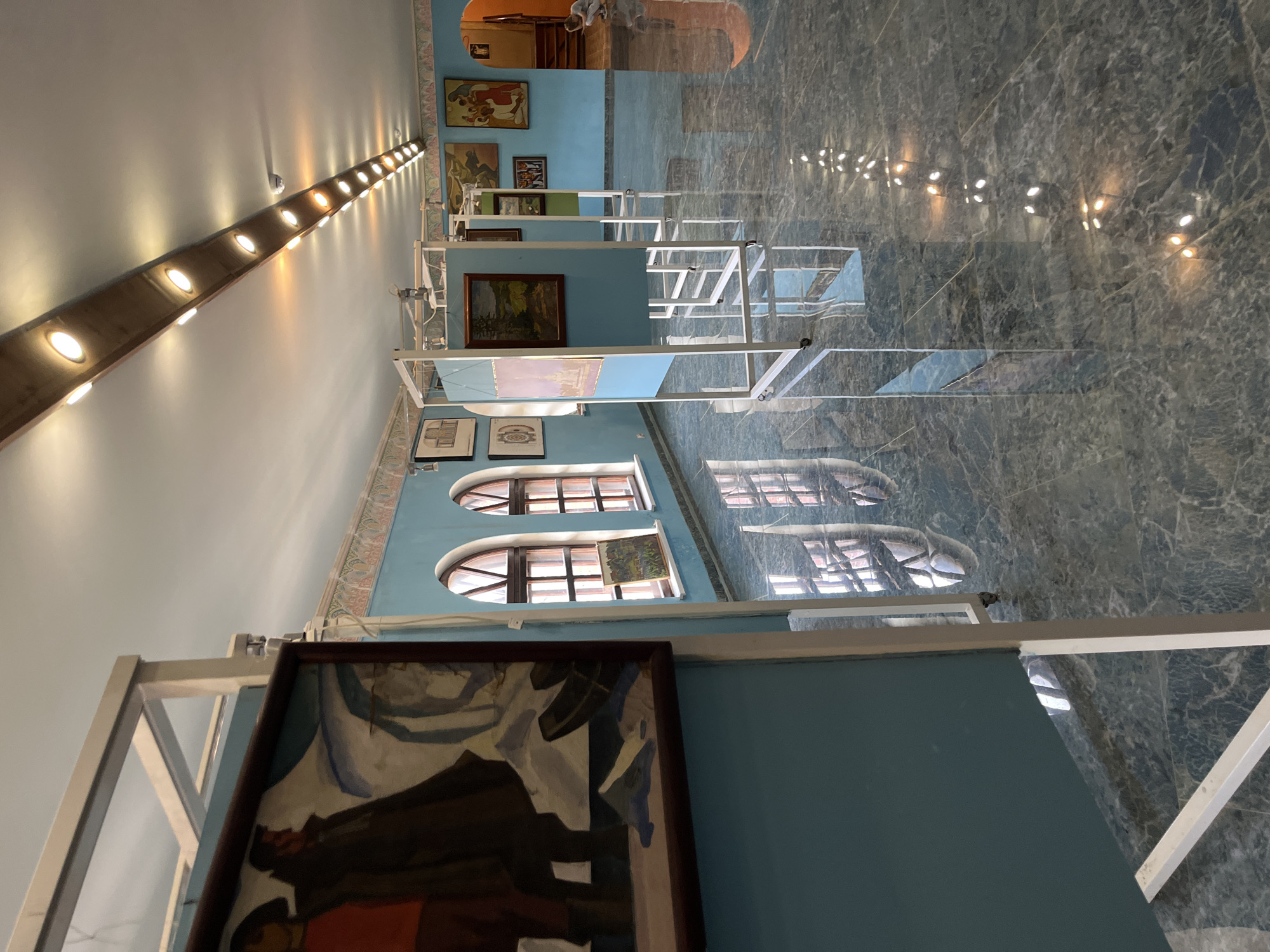 The image depicts an interior art exhibition hall, likely in Kazan, Russia, with a high-gloss, dark marble-patterned floor reflecting the overhead lighting and the displayed artwork. The walls are painted a light blue, and a decorative border runs along the upper section of the wall.
In the foreground, a large framed painting is partially visible, showing two figures in dark coats and red accents against a white and blue abstract background. This painting is displayed on a teal-colored panel supported by a white metal frame.
Further into the hall, several other art pieces are displayed on similar white metal stands, some with teal panels. One painting shows a landscape with trees and a body of water, another appears to be a historical or allegorical scene with multiple figures. There are arched windows on the left wall, revealing a glimpse of the outdoors.
The ceiling features a track of recessed spotlights providing illumination. The floor's reflective surface creates an impression of depth and a mirror-like quality, showing the reflections of the lights and the exhibition structure.
In the background, an archway leads to another area of the building, where a person in light-colored clothing is seen crouching. The overall atmosphere is that of a quiet, well-lit gallery space dedicated to displaying visual art.