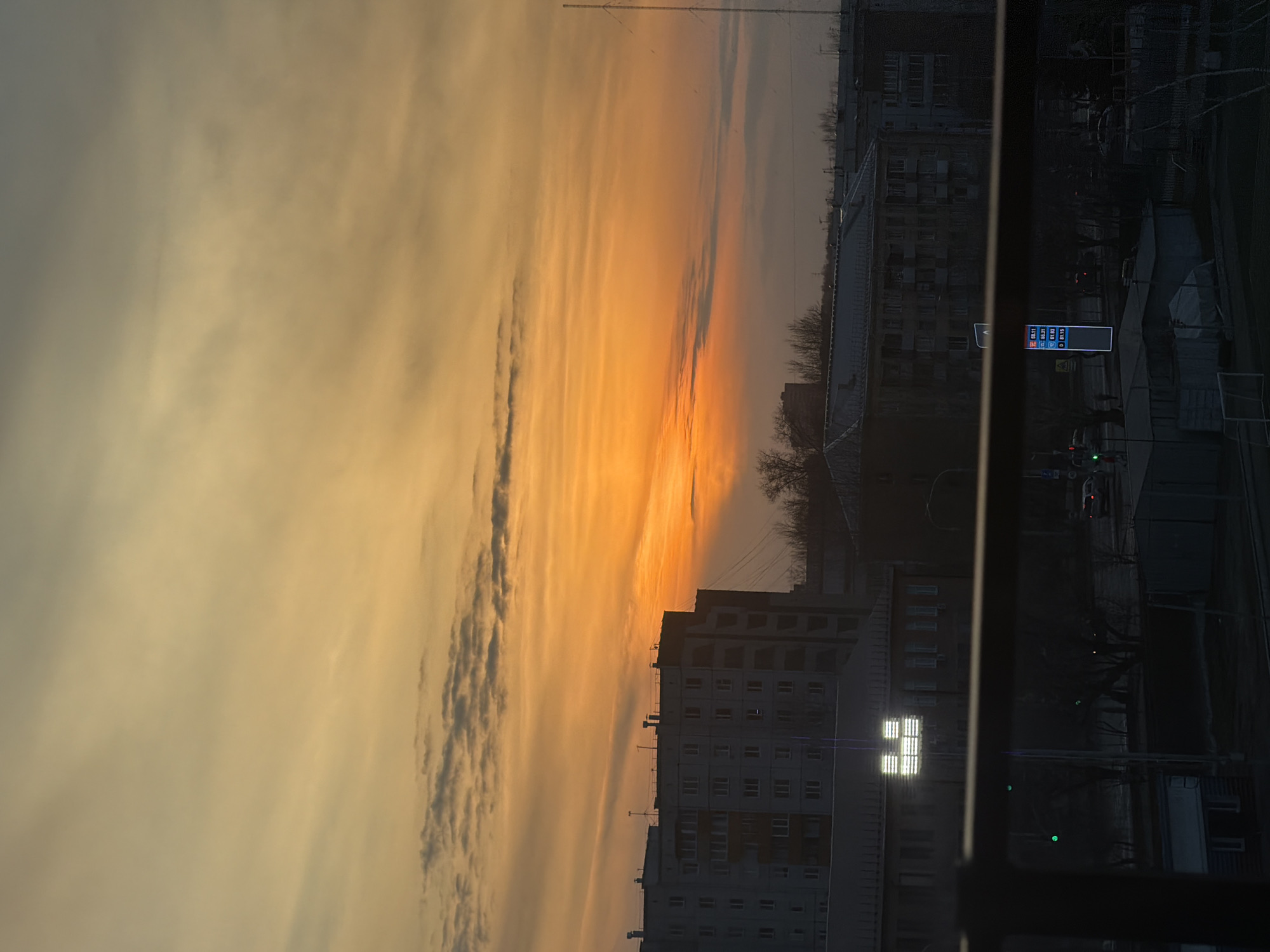 The image depicts an urban sunset or sunrise over Krasnoyarsk, Russia, viewed from an elevated vantage point, possibly a window, indicated by a dark frame on the right. The sky dominates the upper two-thirds of the frame, showcasing a dramatic blend of vibrant orange and yellow hues near the horizon, fading into lighter peach and grey-blue towards the upper left. Long, streaky clouds, arranged horizontally, traverse the sky, enhancing the colorful display.
In the lower part of the image, the cityscape is largely silhouetted against the brilliant sky. Several multi-story apartment buildings, typical of Russian urban architecture, are visible. One building in the mid-ground features a prominent square of bright, illuminated windows. Bare trees, suggesting a cooler season, are scattered throughout the foreground and between buildings. A street with faint vehicle lights runs across the bottom, hinting at ongoing activity.
A tall, slender utility pole or communication tower is faintly visible against the sky on the left. A digital sign near the street displays numerical information, likely fuel prices, which read: "92 45.11," "95 49.31," "98 51.93," and "D 51.15." The low light and striking sky colors create a serene and contemplative atmosphere as day transitions to night or vice-versa.