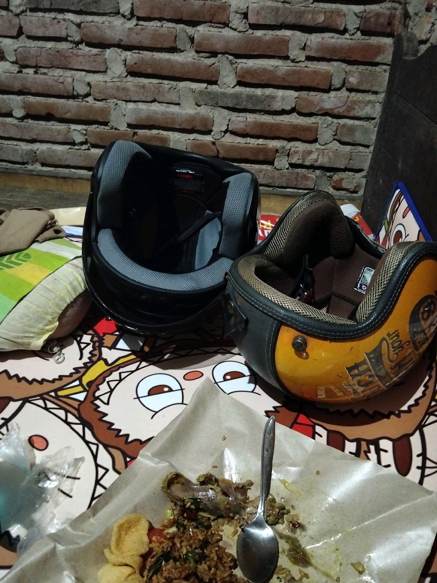 The image shows a close-up view of two motorcycle helmets resting on a surface patterned with cartoon characters. A black helmet with grey lining is on the left, and a yellow and black helmet with some stickers is on the right. In the foreground, a portion of a meal is laid out on brown paper, with a silver spoon resting on it. The background consists of a brick wall. The scene appears to be indoors, possibly a residential setting or a roadside eatery. There are no people visible in the image. The overall lighting is dim, suggesting it might be evening or a poorly lit indoor space. The location is not identifiable as Panimbang, Indonesia, based on the visual information provided.