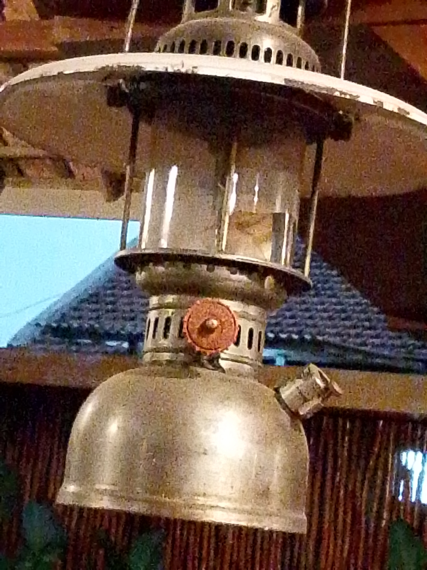 The image shows a close-up of an old-fashioned kerosene lantern hanging from a ceiling. The lantern is made of metal and glass, with a wide brim and a bell-shaped base. A small red knob is visible on the side of the lantern, likely used for adjusting the wick. The background is blurred, but appears to be an indoor setting with wooden walls and a thatched roof visible through a window. The lighting is dim, with the lantern itself appearing to be the primary light source. The scene is static and no people are present. The location is identified as Mojotengah, Indonesia, suggesting this could be in a traditional home or establishment in that region.