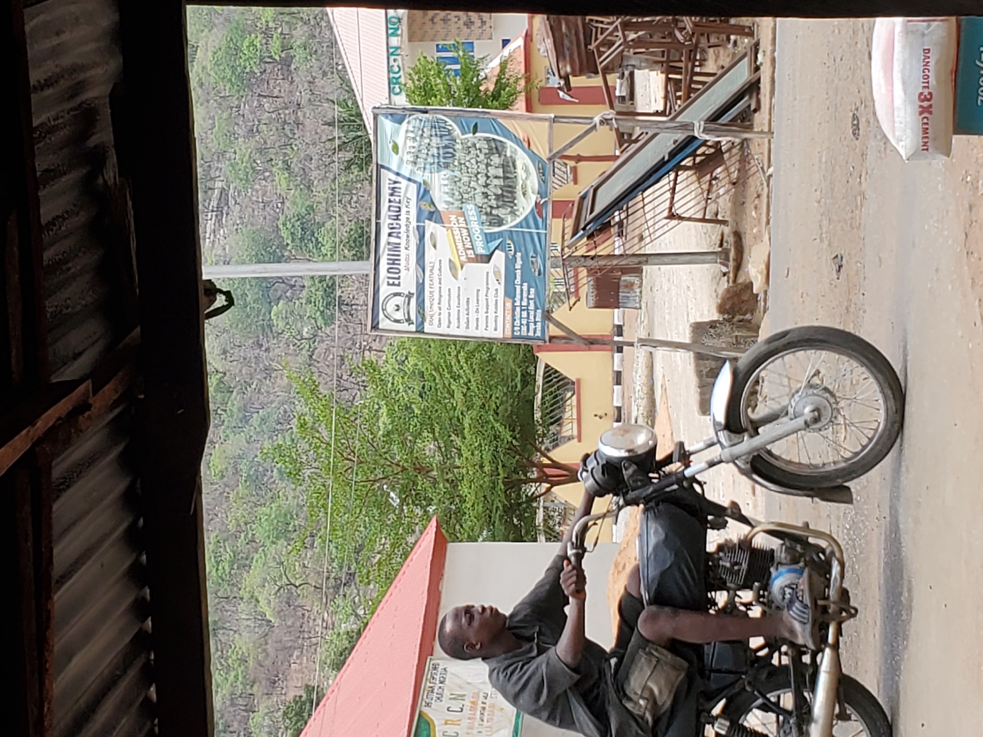 The image shows a man riding a motorcycle on a street in Mararaba, Nigeria. The man is wearing a dark shirt and has a bag slung over his shoulder. He is looking to his left as he drives. The motorcycle is an older model, possibly in need of repair. Behind the man and to the left is a red and white sign. Behind the motorcycle and to the right of the frame, there's a Dangote cement bag and a banner with information about Elohim Academy. The environment suggests a developing area with buildings and a forested mountain in the background.