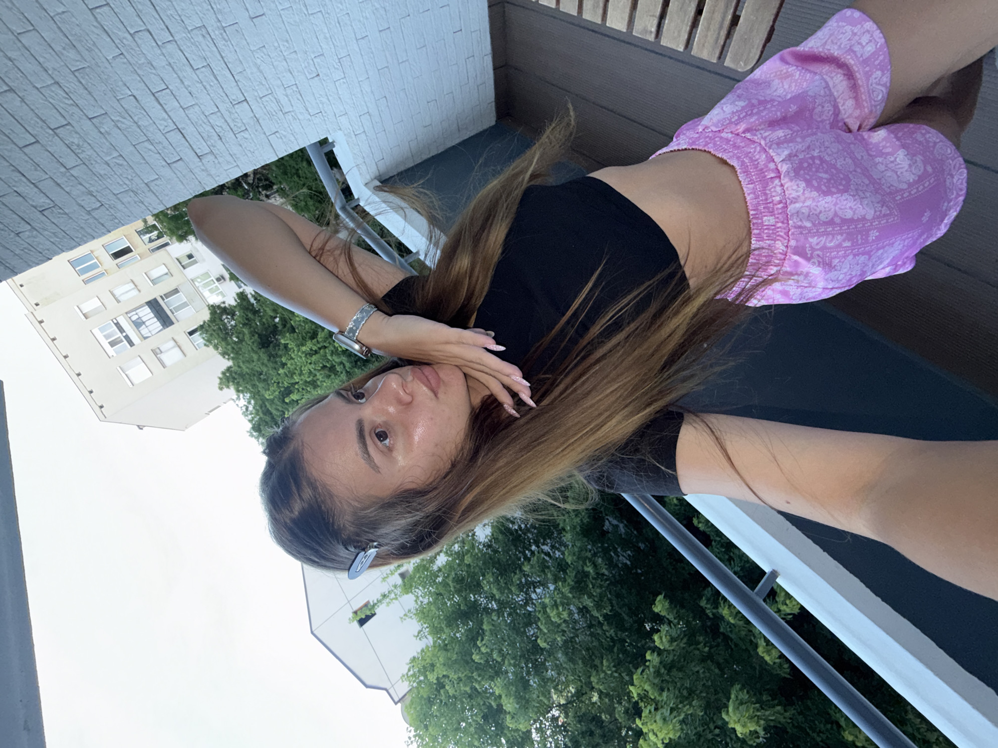 A young woman is captured in a selfie-style shot, lying down outdoors on what appears to be a balcony in Plovdiv, Bulgaria. Her long, light brown hair cascades around her as she looks directly at the camera with a neutral expression. She wears a black cropped top and light pink shorts adorned with a white bandana-like pattern. A silver-toned watch is visible on her left wrist, and her nails are painted a light, possibly glittery, color. Sunglasses rest on her head.
The background reveals elements of an urban setting. To her side and behind her, lush green trees are prominent, contrasting with a multi-story building featuring numerous windows. Above her in the frame, a textured white brick wall or surface is visible, likely part of the balcony or building facade. A balcony railing is also present in the foreground. The bright, clear sky suggests it is daytime, possibly late afternoon or early evening. There is no visible text in the image.