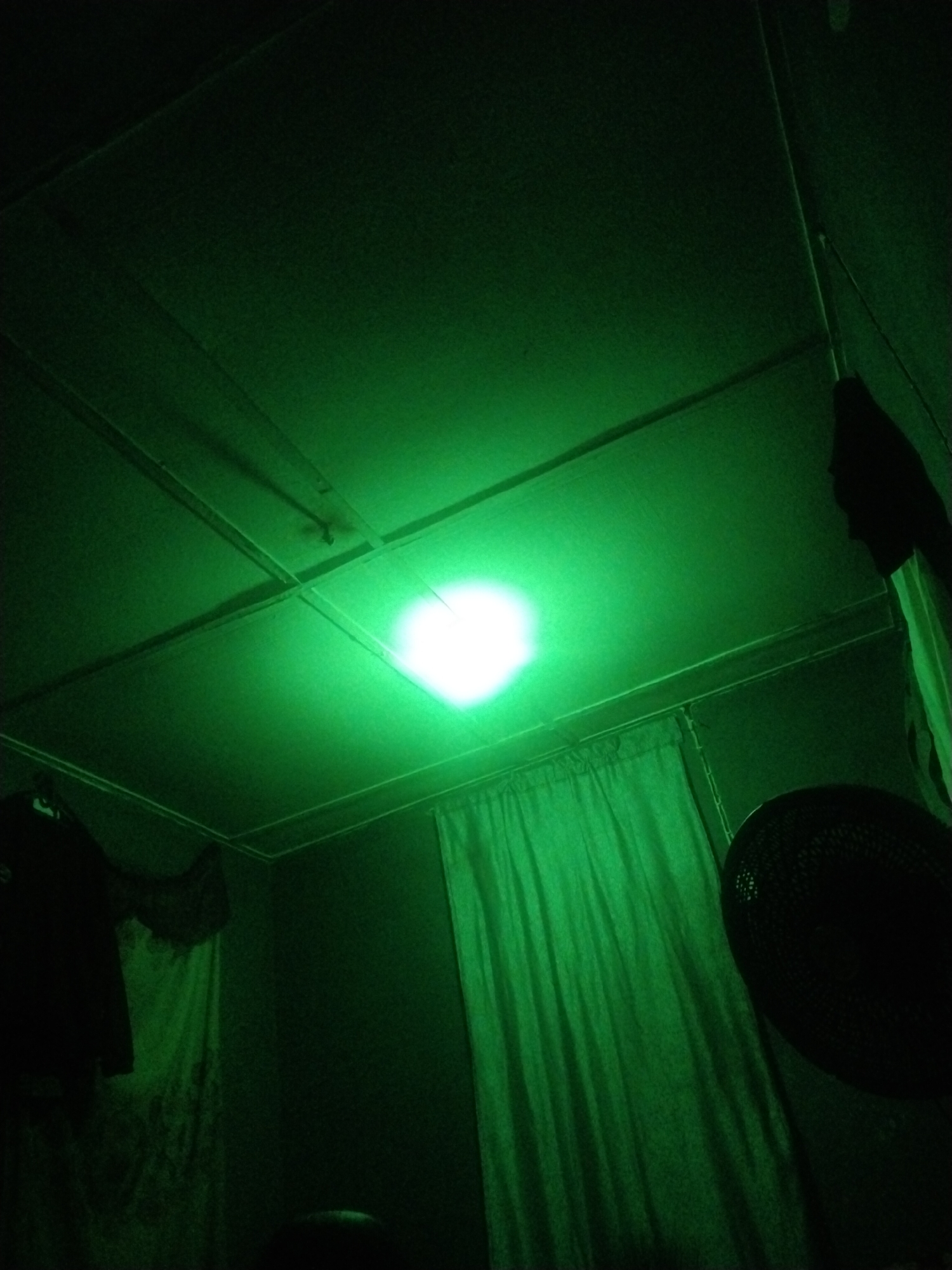 The image shows a dimly lit room with a green hue. The ceiling is composed of rectangular panels. A light fixture, appearing as a bright white circle, is centered on one of the panels, casting a strong green glow. Curtains are draped along one wall, with a dark, circular object, likely a fan, positioned near them. On another wall, a piece of clothing hangs, with a lighter-colored cloth visible behind it. The location is unknown.