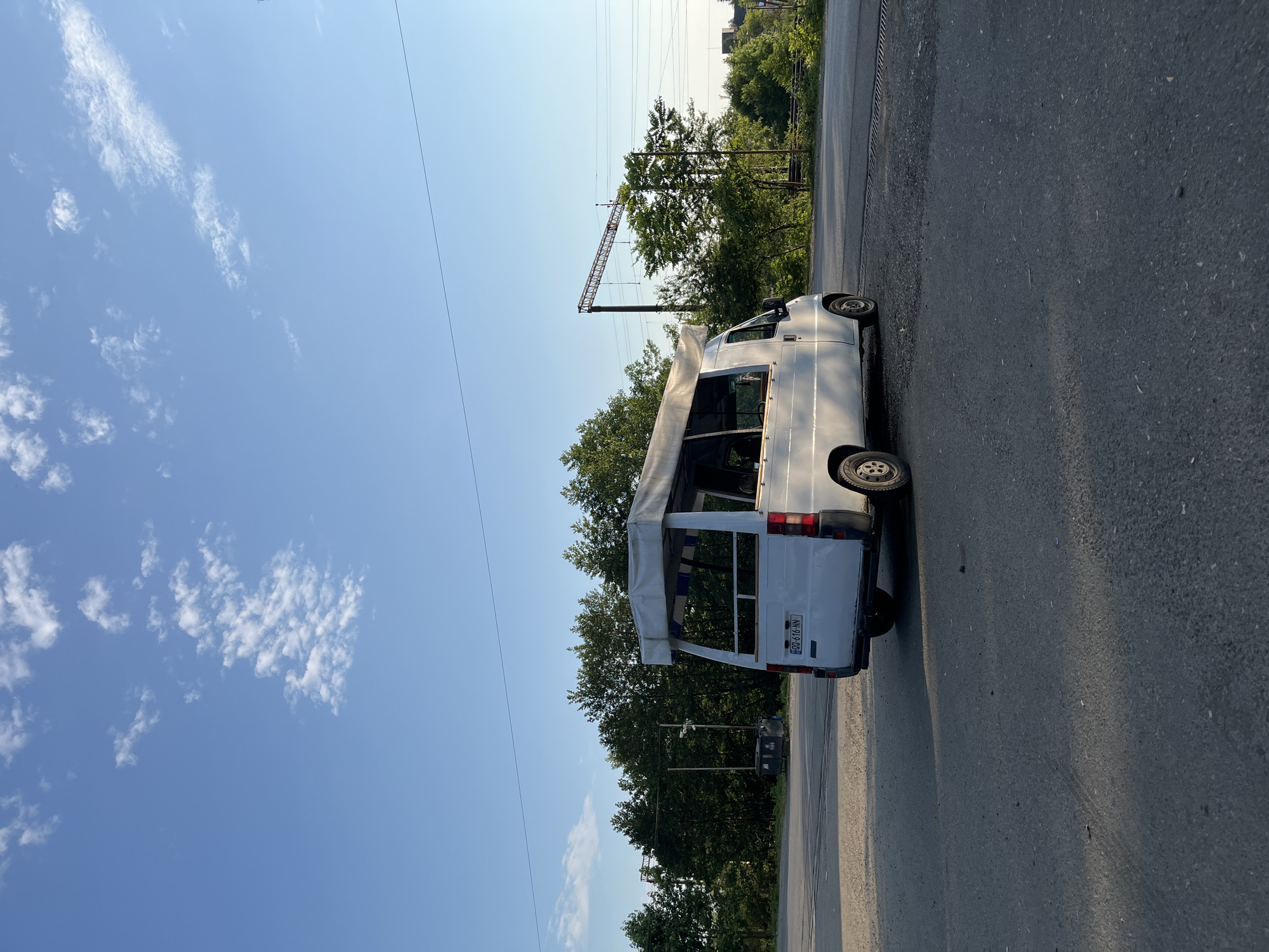 In Ureki, Georgia, a white camper van is parked on an asphalt surface next to a road. The van, viewed from the rear three-quarter angle, has an open canopy extending from its roof. Trees form a dense line behind the van, and power lines and a railway structure are visible in the distance under a blue sky with scattered white clouds. The scene appears to be a roadside area with a clear sky indicating daytime.