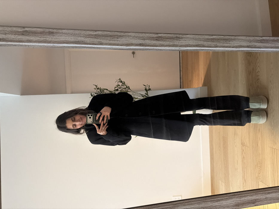 A female subject is depicted taking a full-length mirror selfie in an indoor residential setting, specified as being in the city of New York, United States of America. The subject has dark hair and a faint smile, looking down at a dark-colored smartphone, equipped with a green/olive case, held in both hands. She is wearing a black long-sleeved coat, black flared pants, and light green, thick-soled shoes. The rectangular full-length mirror has a distressed light-colored frame. The background behind the subject's reflection features white walls, a light-colored wooden floor, a white interior door with a silver handle, a white electrical outlet on the left wall, and a green leafy plant positioned behind the subject's right shoulder.