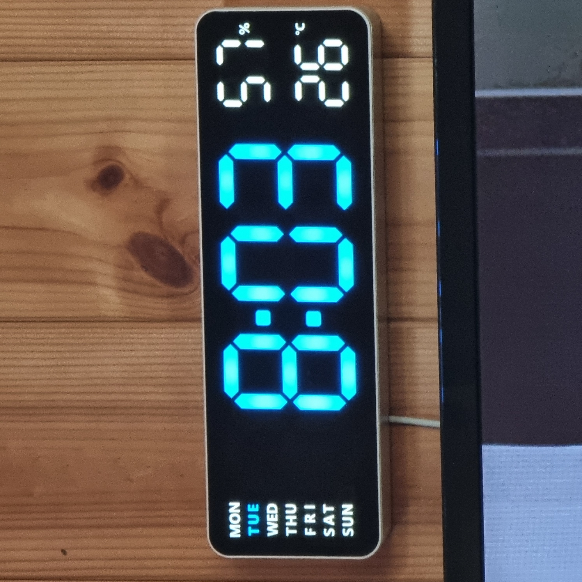 The image shows a digital clock mounted on a wooden wall. The clock displays the time "8:03" in large, bright blue digits. To the left of the time, a vertical list of days of the week is displayed, with "TUE" highlighted in blue, indicating Tuesday. To the right of the time, two readings are shown: "57%" which likely represents humidity, and "26°c" representing the temperature. The clock is a sleek, modern design with a white border and a black display. The wooden wall behind it has a visible grain pattern. Below the clock, the top edge of a dark electronic device, possibly a television or monitor, is visible. The image does not contain any people or indicate a specific location in Dunnae-myeon, South Korea.