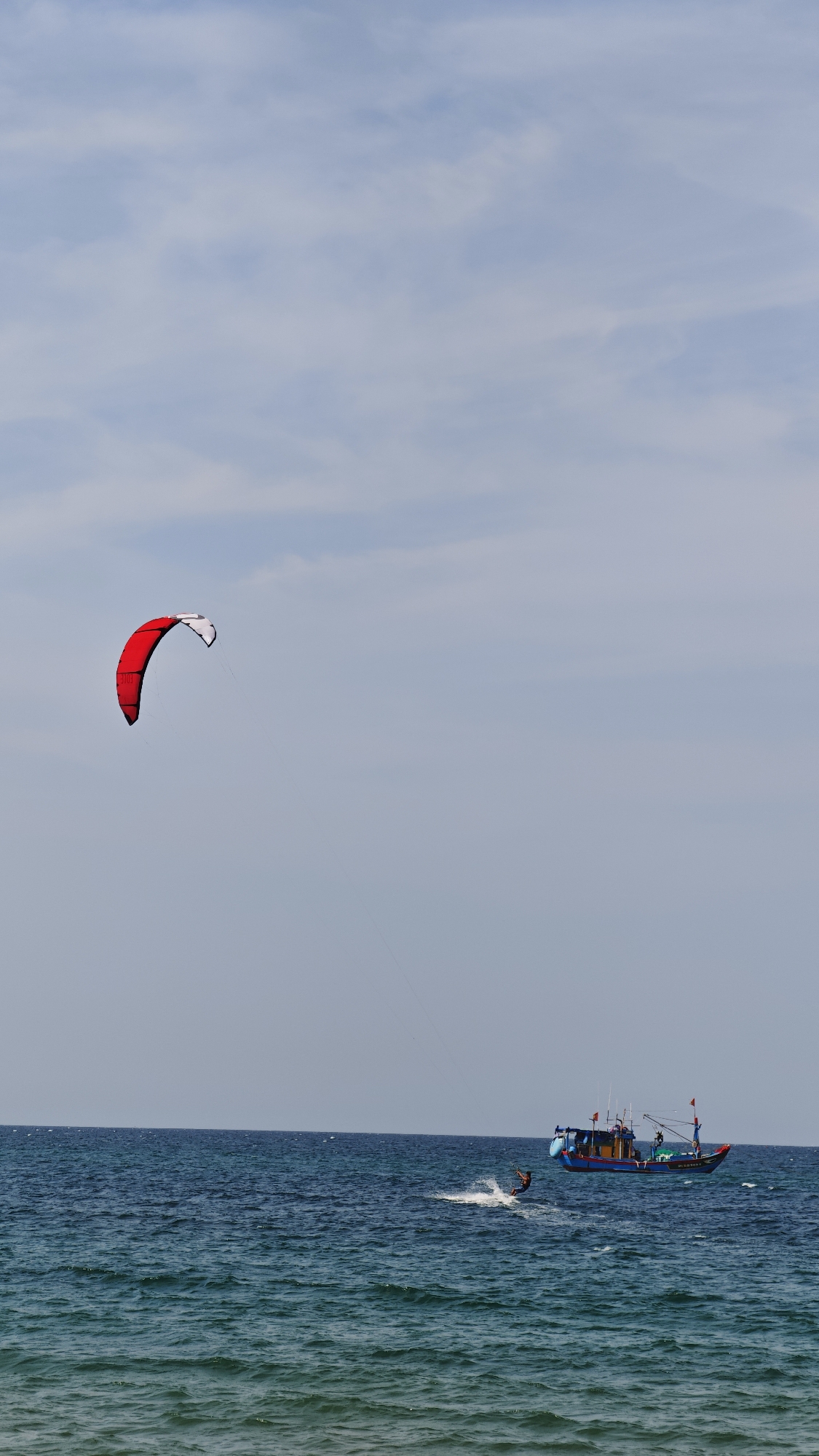A wide shot shows the waters of the ocean in Đà Nẵng, Vietnam. A red and white kite is in the top left of the shot, with a single line extending down towards a kitesurfer below. The kitesurfer is skimming on the water, making a plume of white spray. To the right of the kitesurfer, a small fishing boat is visible with several people on board. The sky is light blue with some clouds.