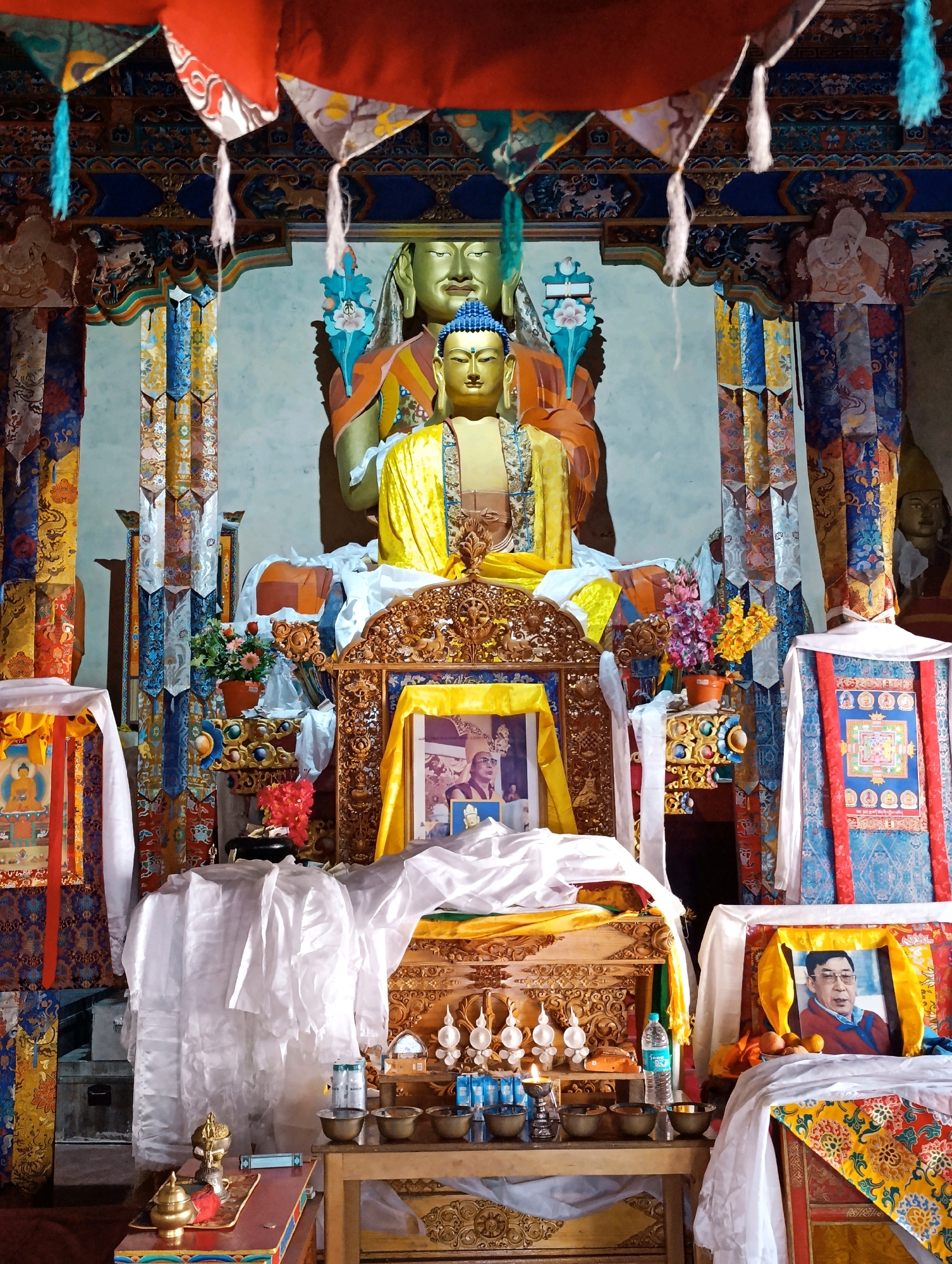 The image depicts the interior of a Buddhist shrine in Pibiting, India. The central focus is a large, golden statue of Buddha, adorned with a yellow robe. Behind it, a larger, golden statue of a Buddha's head is visible. The shrine is elaborately decorated with colorful prayer flags and embroidered banners. Various offerings are placed before the statues, including bowls, bottles of water, and framed photographs. One framed photograph shows a portrait of an elderly man with glasses, wearing a red jacket. Another framed image features a smaller depiction of Buddha. The shrine is illuminated by soft, natural light.