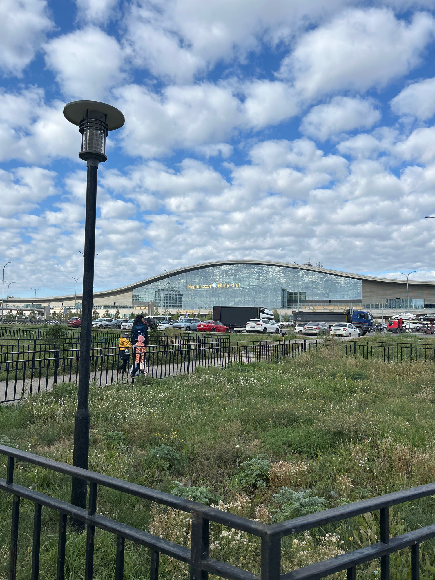 In Astana, Kazakhstan, a modern airport terminal with a curved, glass facade stands under a partly cloudy sky. The text "Нурлы жол" is visible on the building in yellow lettering. In the foreground, a black metal fence encloses a grassy area with scattered wildflowers and a lamppost. Two people, one adult and one child, walk along a paved path behind the fence, with the adult carrying a yellow bag. Various vehicles, including cars and a blue truck, are parked in front of the terminal.