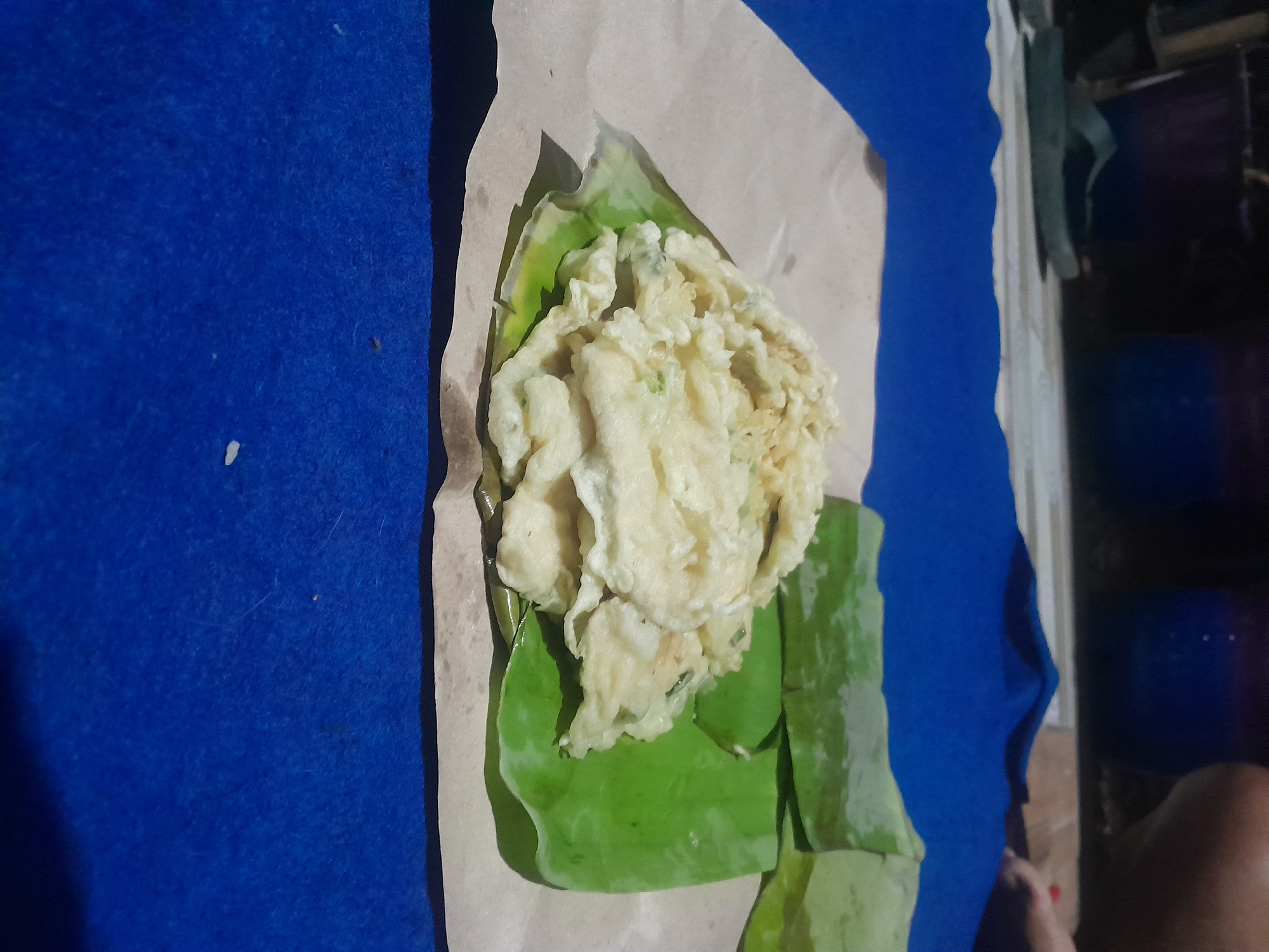 A plate of fried food, likely tempeh, is presented on banana leaves and brown paper on a blue surface. The food is golden brown and appears to be thinly sliced. In the background, out of focus, are several large planters and a glimpse of a person's leg. The overall setting suggests an outdoor or semi-outdoor environment, possibly a market or a casual dining area in Pacul, Indonesia.