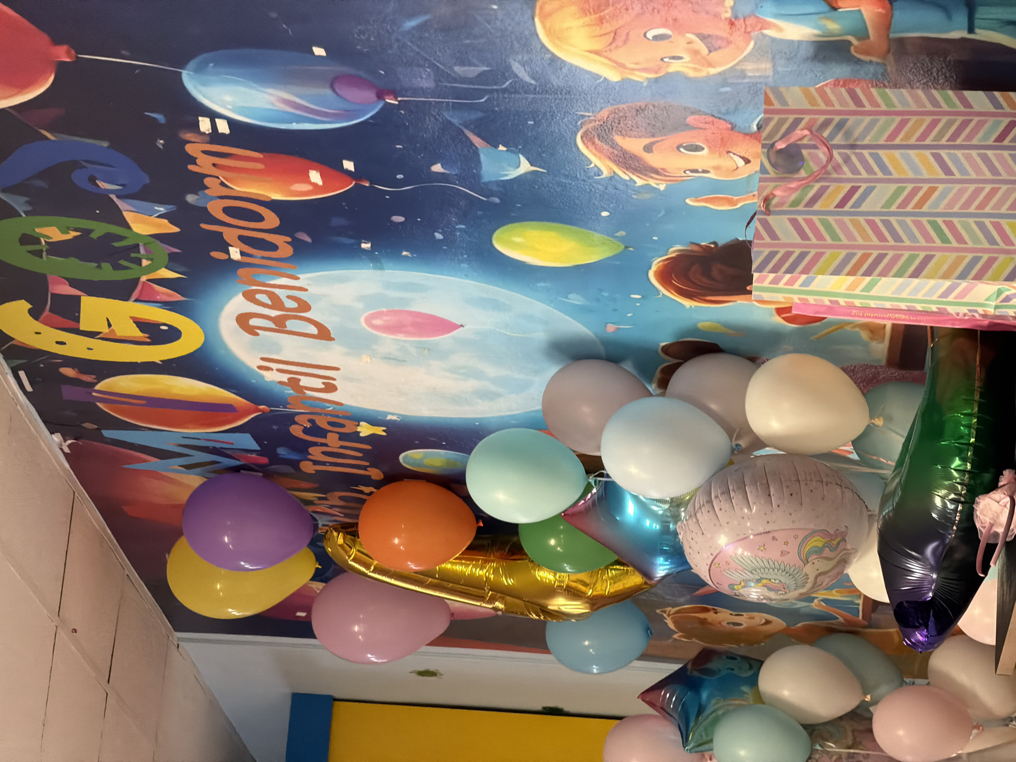 This media file captures a festive scene, likely a children's birthday party, with a prominent display of balloons. The background is a colorful mural depicting cartoon characters, possibly from a children's show or theme, with a celestial or outer space theme. Various balloons are scattered throughout the frame, including round balloons in shades of purple, yellow, pink, light blue, and orange. There's also a large, gold, elongated balloon and a foil balloon with a unicorn design. A patterned gift bag, striped in pink, purple, yellow, and blue, is visible, suggesting gifts were part of the event. The lighting appears to be indoors, possibly from artificial sources, with no indication of time of day or weather. The overall impression is one of joy and celebration. The text "Infantil Benidorm" is visible on the mural, suggesting the event may be related to an event or location in Benidorm, Spain.