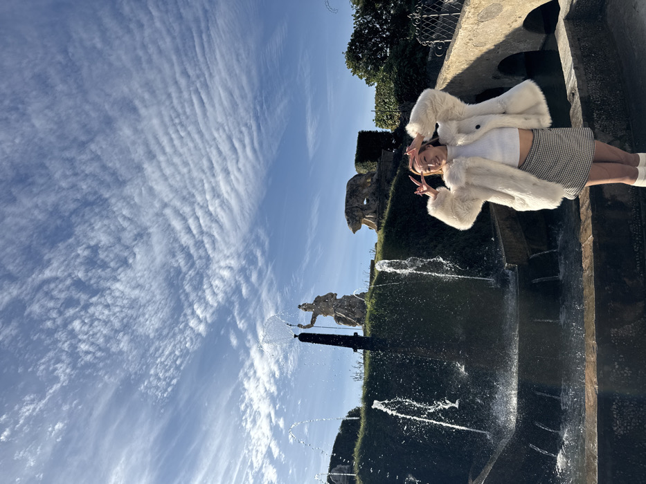 The image captures a young woman posing in the gardens of Villa d'Este in Tivoli, Italy. The woman, situated in the lower right foreground, is smiling at the camera while making a V-sign with both hands, positioned near her temples. She is wearing a cream-colored faux fur coat over a white ribbed crop top, a black and white checkered mini skirt, and white knee-high socks or boots.
Behind the woman, a stone fountain system actively cascades water downwards into multiple tiers and projects several jets upwards. On an elevated green embankment directly behind the fountain, a large stone statue of a classical figure, holding a staff-like object, is visible, from which a distinct stream of water sprays vertically into the air. Further to the right on the same embankment, another stone sculpture depicting a bear is positioned. The background features a bright blue sky dominated by extensive, horizontally arrayed cirrocumulus cloud formations. Stone architectural elements, including an archway, are partially visible on the far right side of the frame. The scene is brightly lit by natural sunlight.