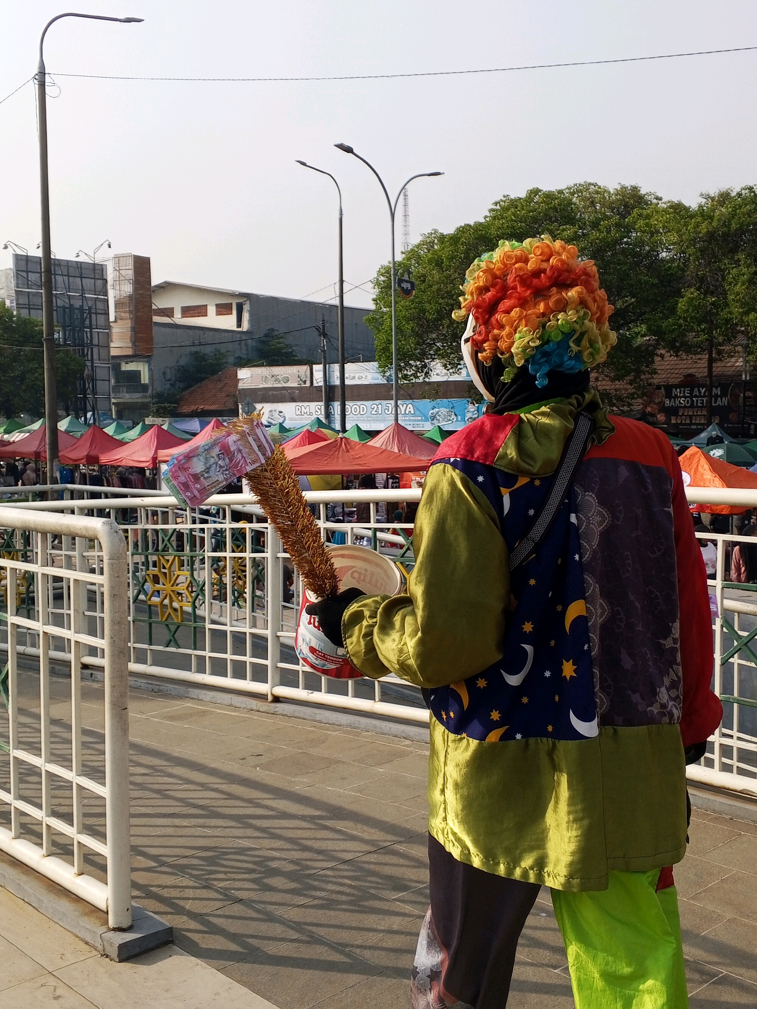 In Serang, Indonesia, a person dressed as a clown stands on a pedestrian walkway, facing away from the camera. They are wearing a vibrant, rainbow-colored wig and a patchwork outfit consisting of gold, red, dark purple, and green fabric. The outfit features a dark blue section with a pattern of yellow stars and white crescent moons. The clown is holding a makeshift collection device in their right hand, which includes a metal can and what appears to be dried plant material with several Indonesian Rupiah banknotes ($100 denominations are visible) attached. The background reveals a bustling market scene with numerous colorful tents, indicating an outdoor marketplace. Buildings and trees are visible in the distance. The walkway is lined with a white metal railing, and the ground is paved with light-colored square tiles. The scene is illuminated by natural daylight, casting distinct shadows from the railing onto the walkway.