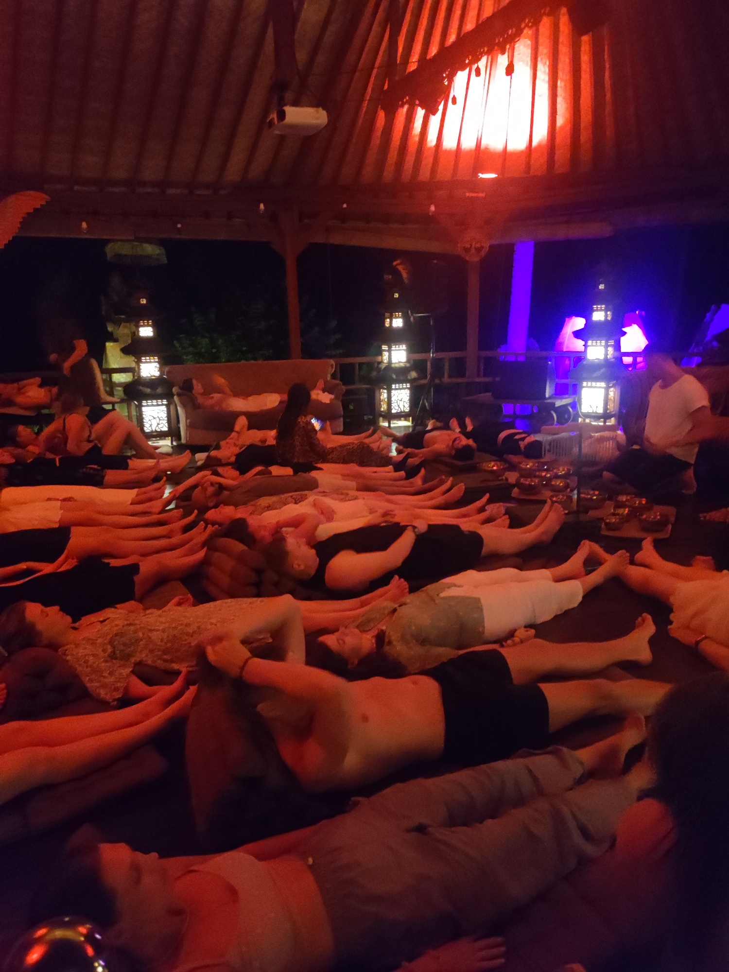 A dimly lit, indoor scene shows a group of approximately 25 people lying on the floor in a large room or open-air pavilion, likely during a relaxation or meditation session. The people are lying supine, with arms at their sides or resting on their chests, and legs extended. A man sits at the end of the row of people, appearing to be leading the session. Numerous small bowls are arranged in front of him. Lanterns decorate the perimeter of the room, and purple and orange lighting sets the mood. A projector is mounted to the ceiling. The environment suggests a spa or retreat setting, possibly in Lokaserena, Indonesia.