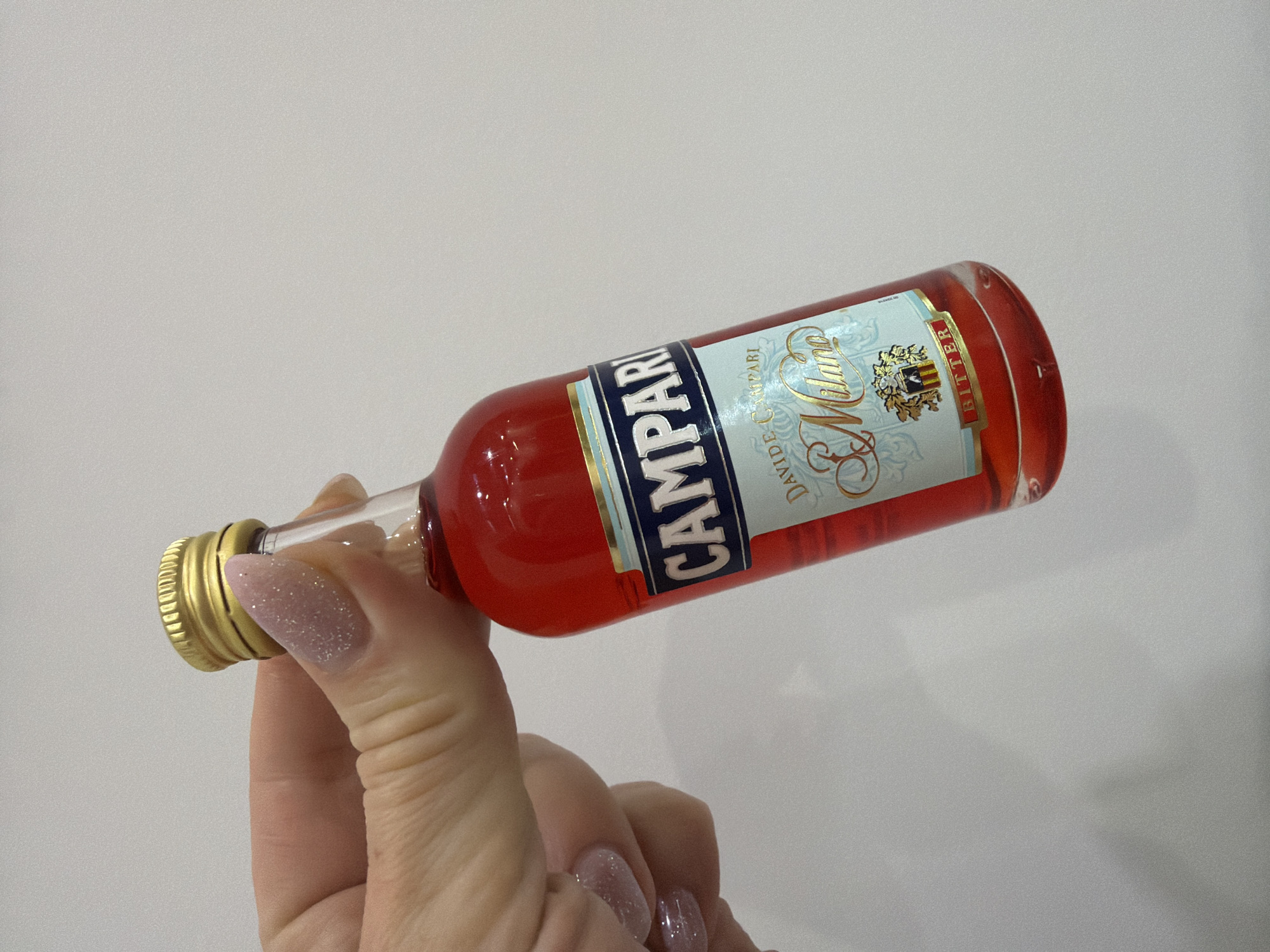 A miniature bottle of Campari liquor, with a red liquid and a gold cap, is held in a person's hand against a plain, light-colored background. The bottle has a label with blue and light blue accents. The label reads "CAMPARI" in large, dark blue letters. Below that, it says "DAVIDE CAMBARI" and then "Milano" in a script font. At the bottom of the label, the word "BITTER" is visible in red letters. The person's fingers, with glitter nail polish, are visible on the left side of the image, holding the bottle. The overall scene is a close-up of the bottle and hand, with no indication of any specific location.