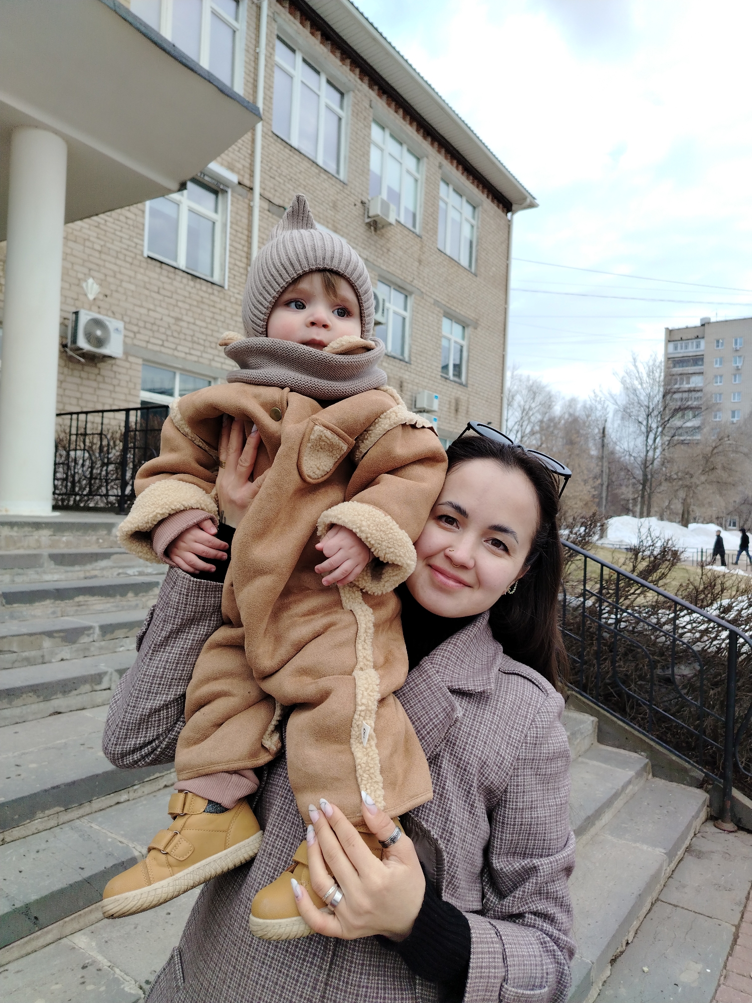 A woman is holding a baby in her arms in front of a brick building. The building has white window frames and an air conditioning unit. The woman is wearing a plaid coat and has sunglasses on her head. The baby is wearing a brown jumpsuit with fur trim and a knit hat. They are standing on a set of grey concrete stairs with a black iron railing. In the background, there is a taller apartment building and trees. There is snow on the ground. People are walking in the background. Based on buildings in the background, this image was taken in the city of Chaikovsky, Russia.
