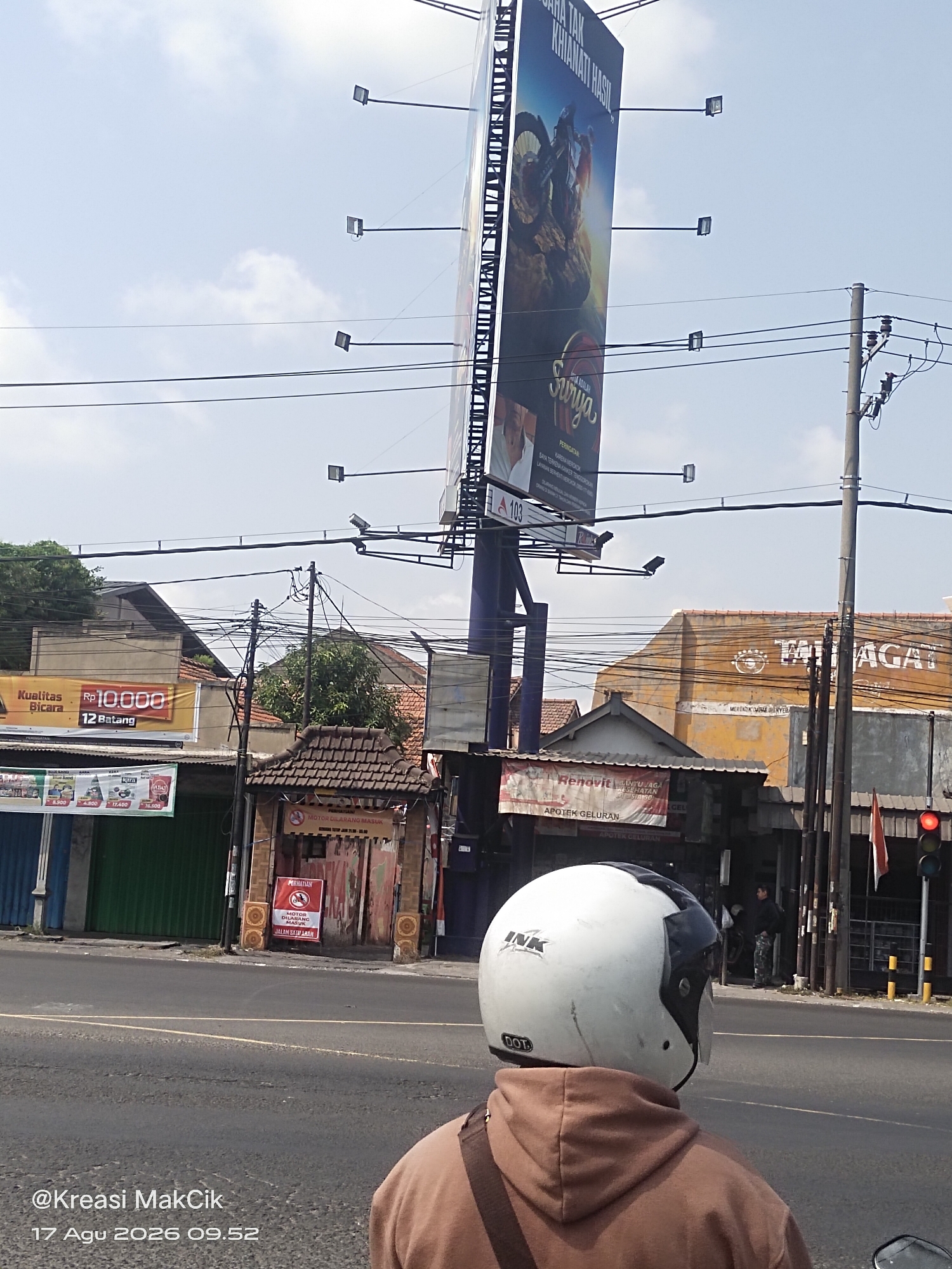 This is a street-level, eye-level, full-shot photograph taken on a clear day in Kalijaten, Indonesia. The foreground is dominated by the back of a person wearing a brown hooded sweatshirt and a white "INK" brand motorcycle helmet with "DOT" visible on the back. The person appears to be on a motorcycle, indicated by the reflection of a mirror on the bottom right.
The middle ground of the image features a street with a double yellow line and a white dividing line, suggesting a multi-lane road. To the left of the street, there are various shops and buildings. One building has a prominent banner that reads "Kualitas Bicara Rp 10.000 12 Batang". Another banner advertises "Renovit BANTU JAGA KESEHATAN". Further down, a small building with a tiled roof has signage that says "MOTOR DILARANG MASUK" (Motorcycles are prohibited) above a red circular sign with a motorcycle icon crossed out.
In the background, a massive billboard towers over the scene, displaying an advertisement for "SURYA" brand cigarettes. The billboard features an image of a person on a motocross bike and text in Indonesian. To the right of the billboard, a building with orange walls has large white lettering that reads "TATAGAT". Utility poles with numerous tangled wires are visible throughout the scene. A traffic light with a red light illuminated is present on the far right. The sky is bright blue with scattered white clouds. The timestamp "@Kreasi MakCik 17 Agu 2026 09.52" is visible in the bottom left corner of the image.