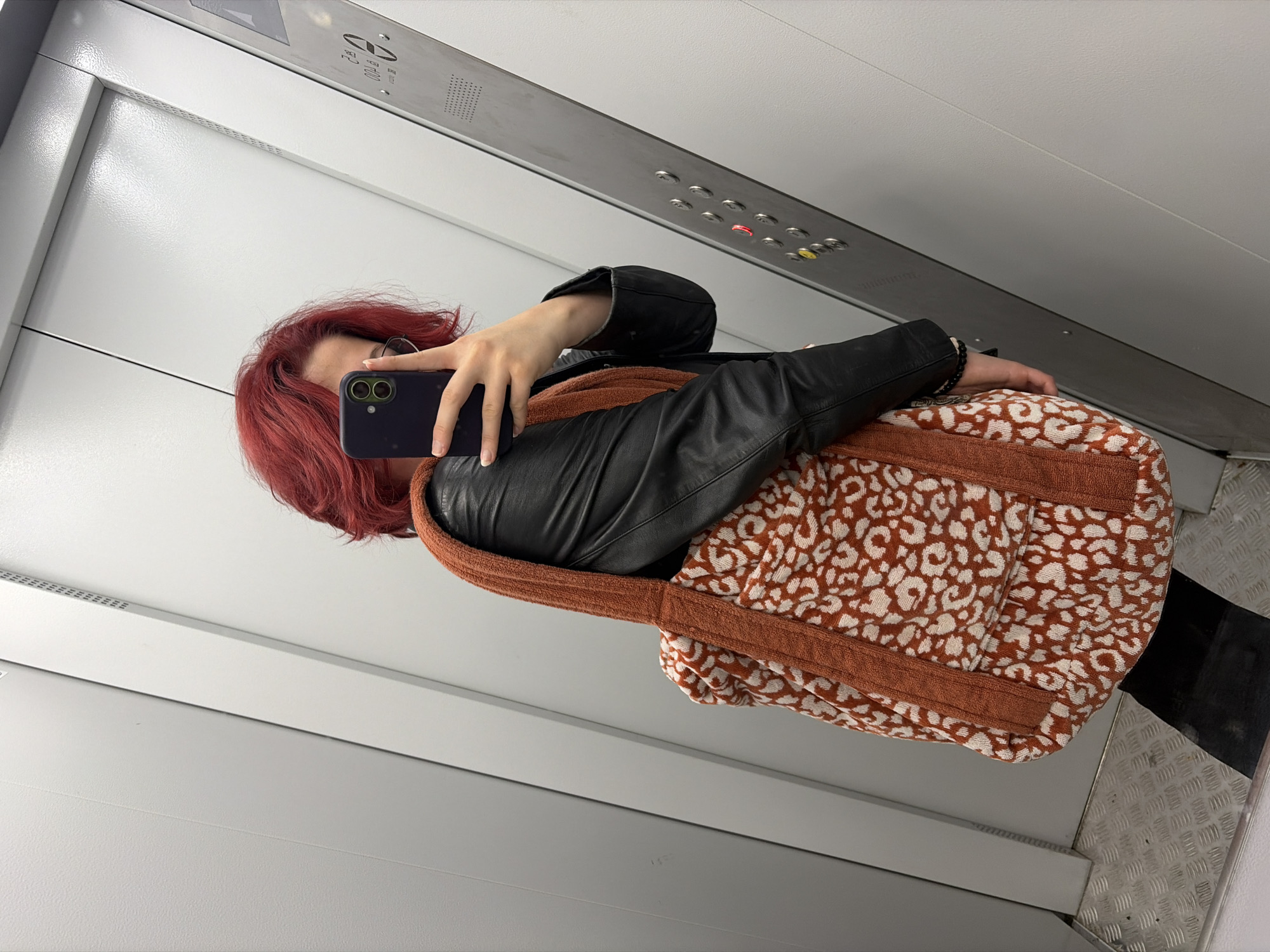 An individual with vibrant red hair, wearing a black leather jacket, holds a smartphone to take a mirror selfie inside an elevator. The person is carrying a large tote bag with a distinctive orange and white leopard print pattern. The elevator interior features metallic walls, an elevator control panel with illuminated buttons, and a textured metal floor. The background suggests an urban setting, possibly within a building in Chelyabinsk, Russia, given the commonality of such elevators in Russian cities.
