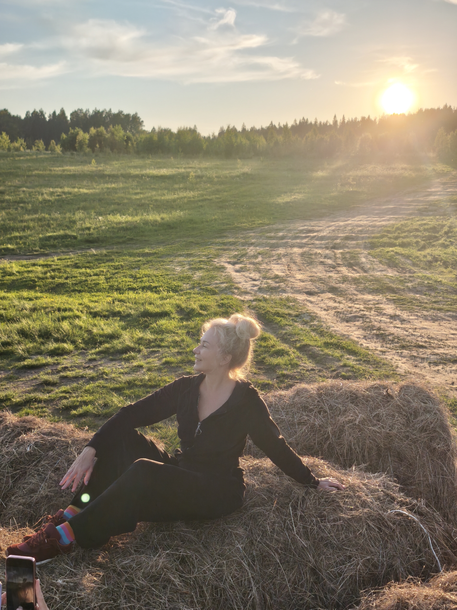 This vertical image captures a serene outdoor scene during the golden hour, likely in Стеклянный, Russia, as the sun sets. The warm, golden light of the low sun emanates from the upper right, casting a strong lens flare and illuminating the landscape.
The main subject is a woman, appearing middle-aged, with blonde hair styled in a messy bun. She is sitting comfortably on a large, round bale of hay, positioned towards the lower center of the frame. She wears a dark, possibly black or dark brown, long-sleeved jumpsuit or tracksuit, accented by vibrant, multi-colored striped socks and dark sneakers. Her head is turned to the left, and she has a gentle, contented smile on her face, suggesting a relaxed and happy mood.
The setting is an open, rural field, characterized by patches of green grass and bare earth or dirt paths. A dense line of trees forming a forest marks the horizon in the background, under a partly cloudy sky. The low angle of the sun creates long shadows and highlights the textures of the hay bale and the field.
In the bottom left corner, a smartphone held by another individual is partially visible, indicating that the woman is posing for a photograph. The overall atmosphere is peaceful and natural, showcasing a tranquil moment at sunset. No visible text is present in the image.