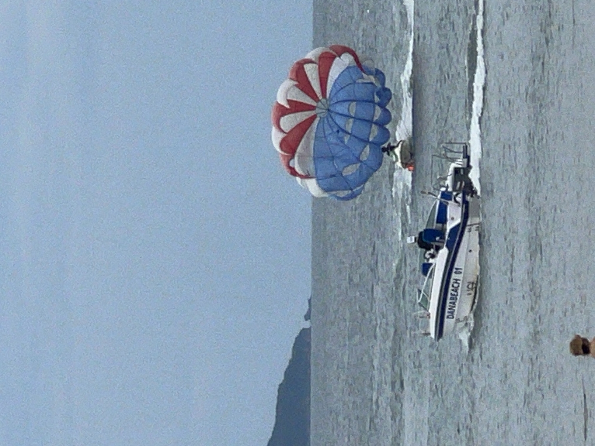 A parasailing operation is underway in the ocean off the coast of Da Nang, Vietnam. A speed boat labeled "DANABEACH 01" is moving from left to right across the frame, creating a wake. In the distance, another boat is pulling a person on a parasail. The parasail is a large, circular canopy with red and blue sections. The sea is choppy under an overcast sky. In the far distance, a dark, hilly landmass is visible on the horizon. In the foreground, a person's back and head, wearing a hat, are visible near the bottom of the frame, suggesting they are on the shore.