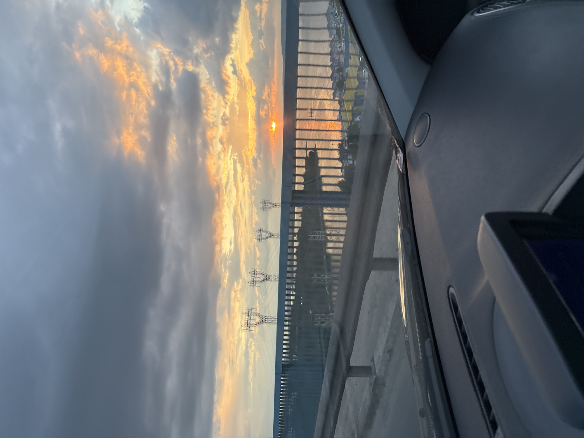 The image shows a sunset view from inside a vehicle, likely a car, crossing a bridge or elevated roadway. The location is identifiable as Varna, Bulgaria, based on the presence of a distinctive series of overhead high-voltage power lines running parallel to the road and the view of the harbor or coastline. The sun is setting over the water, creating a bright orange glow in the clouds. The guardrail of the bridge is visible through the windshield. Part of the interior of the car, including the dashboard and infotainment screen, is visible in the foreground. No people are clearly visible, though a blurry silhouette might be visible through the windshield on the bridge.
