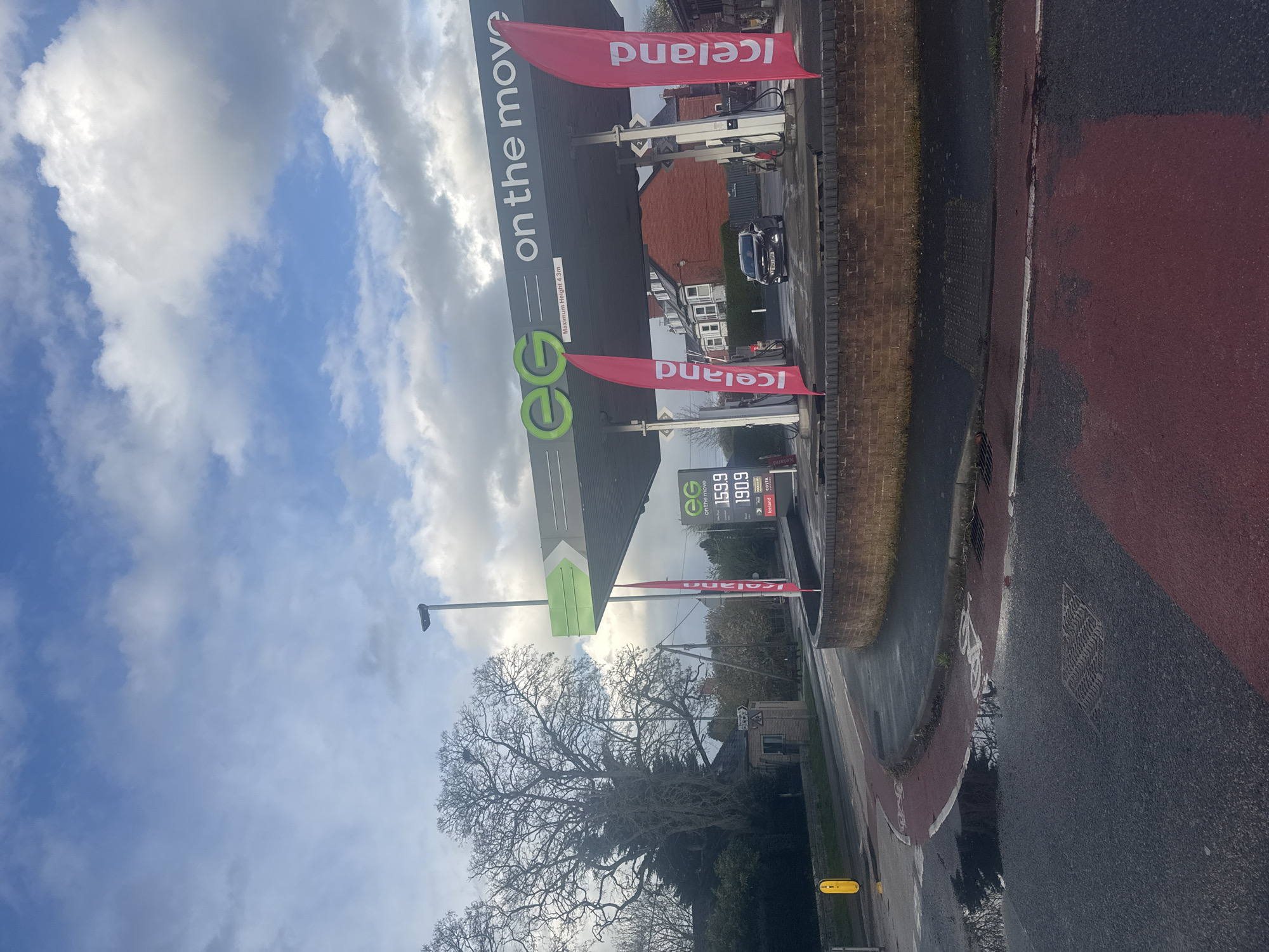 This outdoor shot is taken in Welshpool, United Kingdom, focusing on an EG fuel station with Iceland food market affiliation. The fuel price display shows gasoline at 159.9 and diesel at 190.9. "Iceland" flags flank the EG-branded fuel station canopy. The sky is partly cloudy. Street furniture, including a lamppost and traffic control equipment, are present. A brick wall borders a red-marked pavement and road surface. A large tree with bare branches is positioned in the foreground.