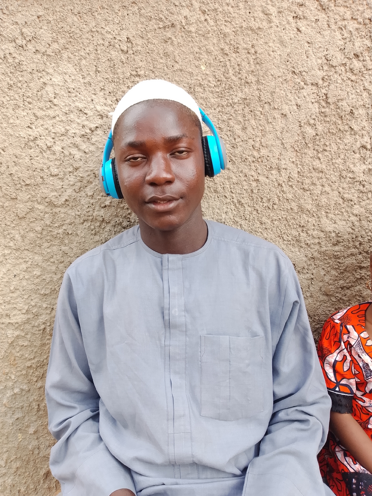 A young Black male wearing a white kufi and a light blue, long-sleeved traditional shirt is seated against a textured, light brown wall. He is wearing bright blue headphones over his ears and looking towards the camera with his eyes slightly squinted. To his right, a portion of another person wearing a vibrant orange and red patterned shirt is visible. The overall scene appears to be outdoors, likely in a public or semi-public space within the city of Jos, Nigeria, given the architectural style of the wall.