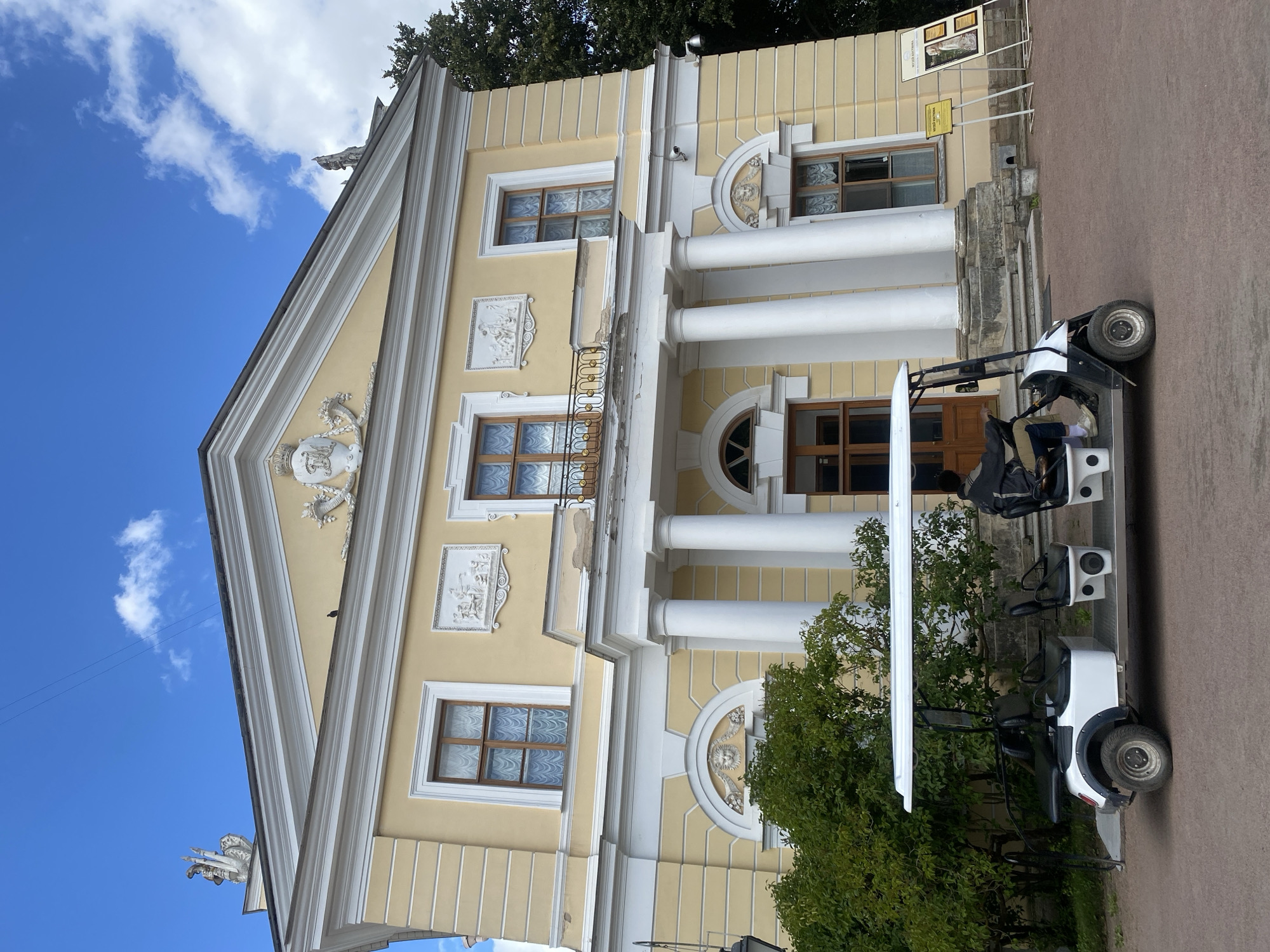 In Pavlovsk, Russia, a yellow neoclassical building with white columns and decorative reliefs stands under a blue sky with scattered clouds. In the foreground, a white electric vehicle, similar to a golf cart, is parked on a reddish-brown path. A person wearing a dark jacket and beige pants is seated in the driver's seat of the vehicle. The building features multiple windows with ornate frames and white curtains. Green trees are visible to the left and right of the building, and a display board with images and text is situated to the right.