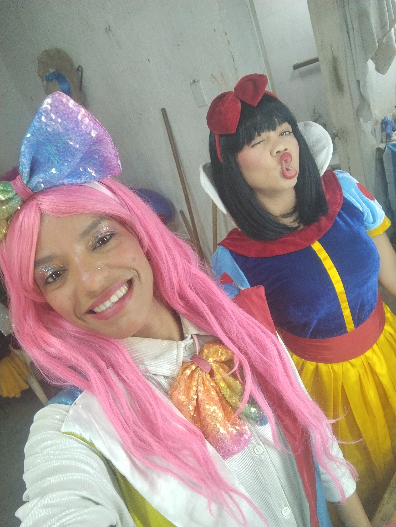 Two women are posing in a brightly lit, likely indoor, location. One woman is in the foreground, wearing a bright pink wig, a sequined hairbow of light blue, purple, and green, a white and iridescent jacket, and a large sequined bowtie with various colors. She is smiling and has glitter-like decorations around her eyes. The other woman is dressed as Snow White, with black hair, a red bow in her hair, and the iconic blue, yellow, and red Snow White dress with a white collar. She is winking and pouting her lips. The background is cluttered with props and clothing, suggesting a backstage area. The scene is likely in Porlamar, Venezuela, as specified in the instructions. No particular city is evident based solely on the visual evidence.
