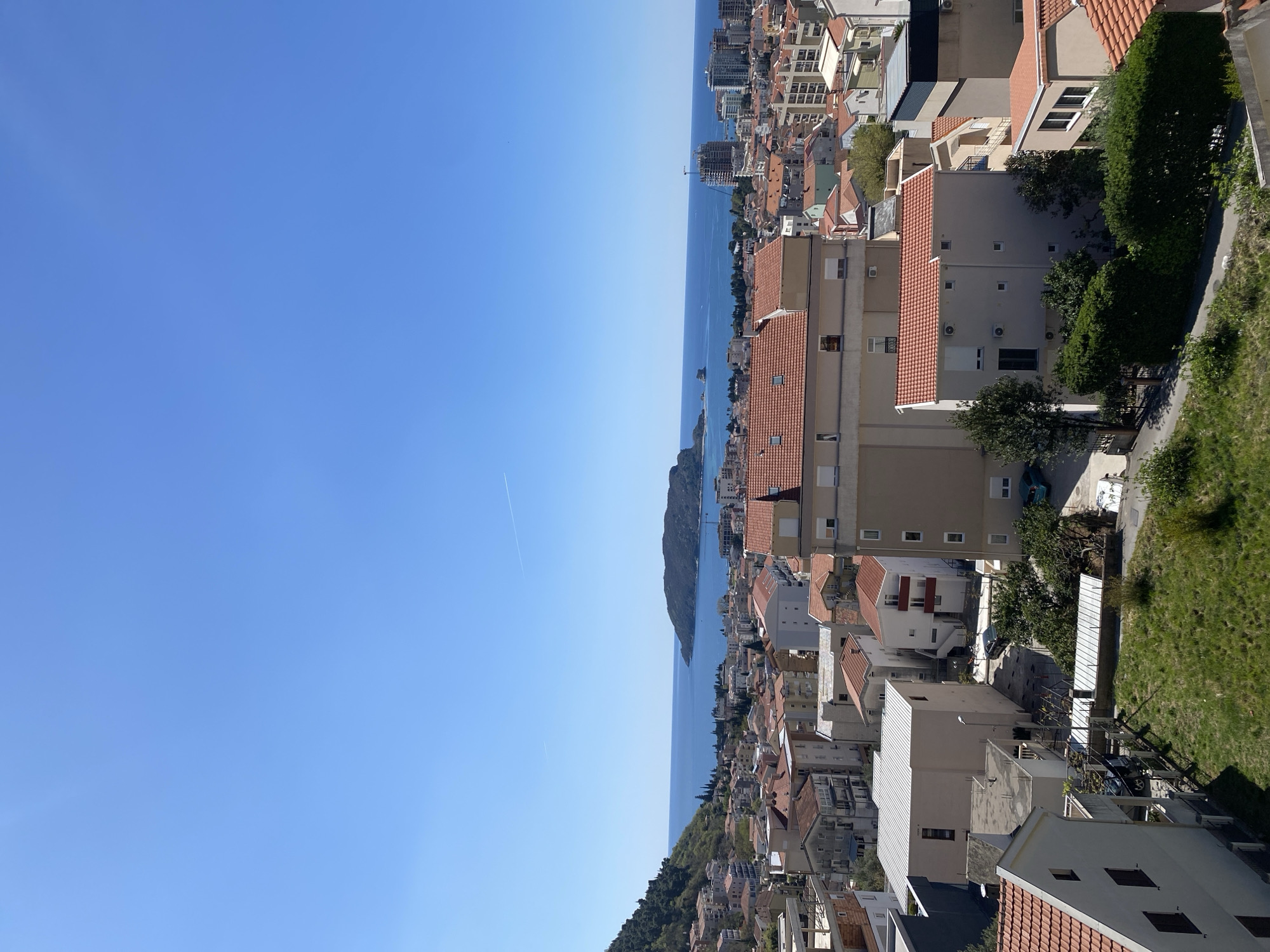 The image is a high-angle view of Budva, Montenegro, encompassing the cityscape, coastline, and clear blue sky. The city is composed of closely packed buildings, predominantly residential, with orange tile roofs. Buildings vary in height, with some taller structures near the coast. The Adriatic Sea stretches along the middle of the frame, bordered by the urban area on one side and the clear sky on the other. A mountainous peninsula is visible in the sea. Vegetation, including trees and shrubs, is interspersed among the buildings and along the coastal edge. A contrail is visible in the sky. No people are visible in the image.