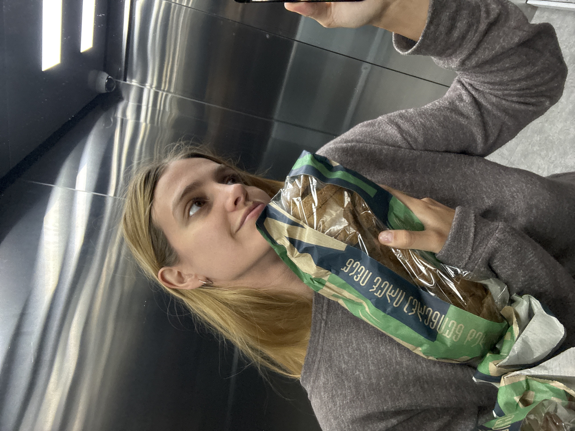A young woman is captured in a selfie, with the image rotated 90 degrees counter-clockwise. She has light skin and long, straight blonde hair, parted on the side. Her head is tilted slightly, and she looks upwards with a neutral expression. She wears a grey, long-sleeved top with a visible sleeve texture.
In her right hand, she holds a smartphone, likely used to take the photograph. With her left arm, she holds a clear plastic bag containing several pieces of what appears to be dark bread or pastries. The bag is partially covered by a decorative paper sleeve featuring blue and green stripes. Visible text on the sleeve is in Georgian script, reading "ფფუ ფქვ" and another line starting with "კეყტ" and ending with "ყფ".
The background features reflective, metallic walls, characteristic of an elevator or a modern, industrial interior. Bright rectangular light fixtures are visible on the ceiling, casting reflections on the metallic surfaces. In the upper left corner of the image, a small, black security camera is mounted in the corner of the room. The overall setting is indoors.