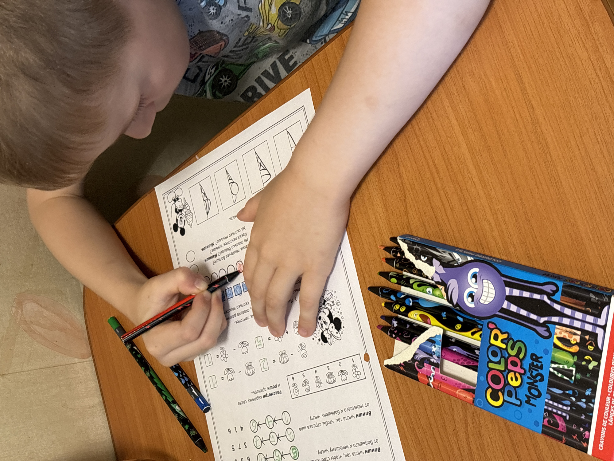 The image depicts a child seated at a brown wooden table, engaged in a worksheet. The child, with fair skin and light brown hair, is wearing a gray shirt with "RIVE" printed on it. They are focused on coloring or marking the worksheet with a red and black pen.
The worksheet contains math problems and images. A box of "Color Peps Monster" colored pencils sits to the right of the worksheet. Two additional pens, one green and black and one blue and black, lie near the child's left hand.
A partially erased marking is visible on the left side of the table near the edge.
There is no information about the location Rusanovo, Russia, in the image.