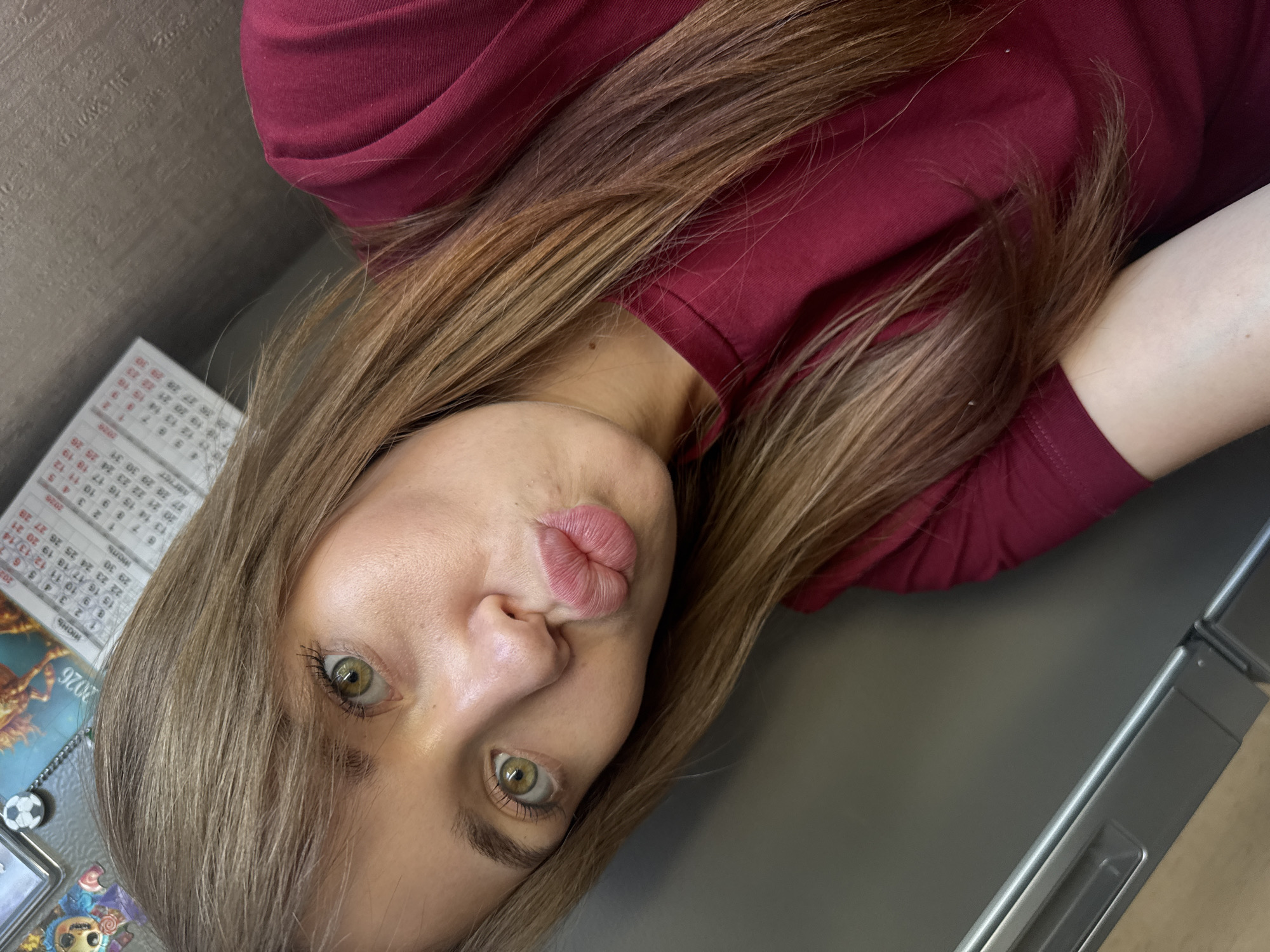 A young woman is taking a selfie, holding the camera at an angle that captures her face and upper body. She is wearing a maroon long-sleeved shirt. Her long, brown hair is falling around her face and shoulders. Her lips are pursed, giving a playful expression. Her eyes are a striking green. In the background, a calendar with the year "2026" visible is attached to a metallic surface, possibly a refrigerator or a locker. The calendar displays text in Russian, indicating the month "Июнь" (June). There are also some decorative magnets on the surface. The lighting appears to be indoor, with no indication of the time of day or weather conditions. The overall impression is a casual, personal moment captured for a social media post or personal memory.