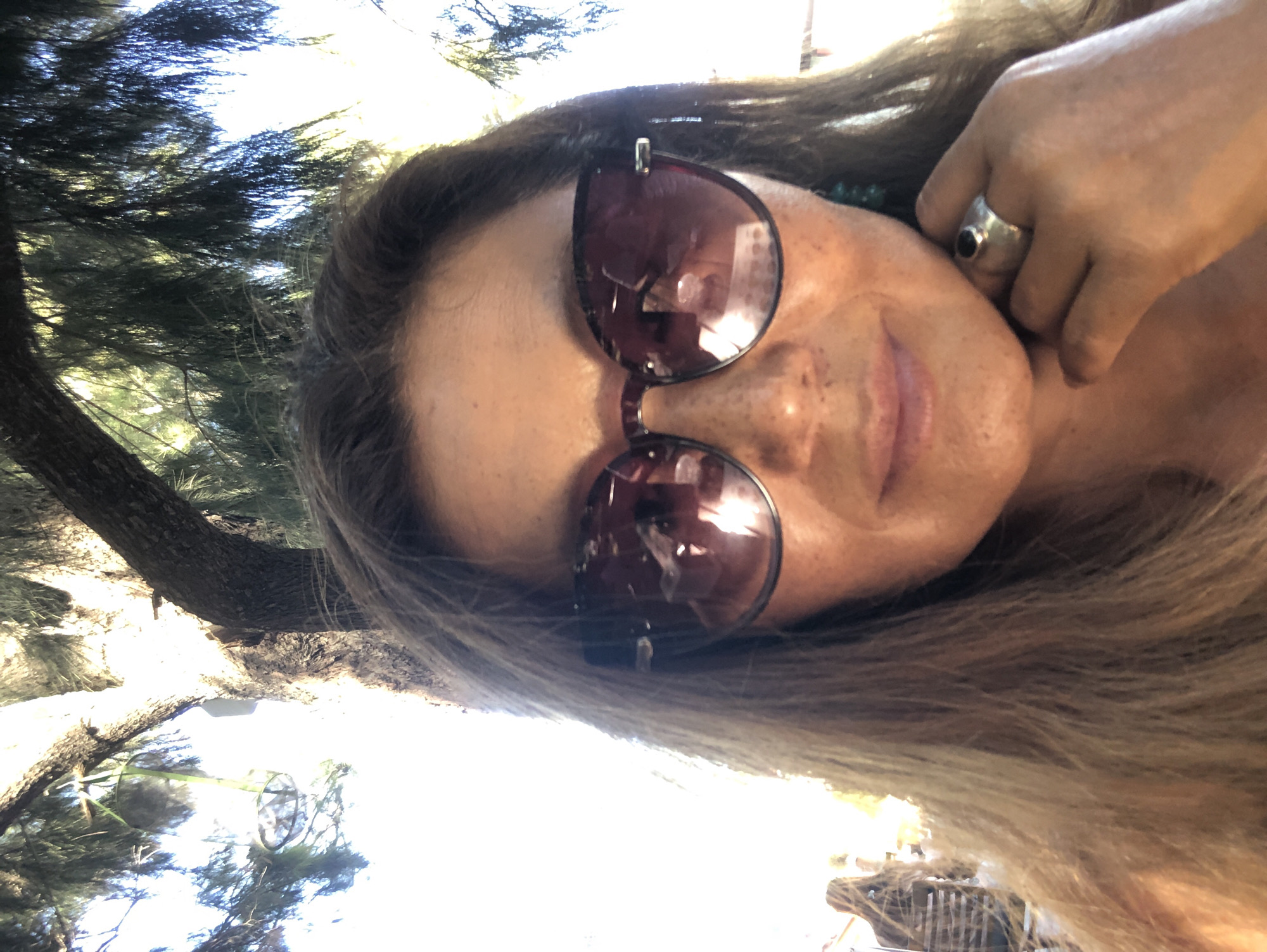 A woman takes a selfie outdoors, partially obscured by trees and foliage. She is wearing large, dark-rimmed sunglasses with reflective lenses and a silver ring with a dark stone on her right index finger. Her hand rests near her chin. Her hair is long and brown, with some strands falling across her face. The background suggests a natural environment with dappled sunlight filtering through the leaves. There is a hint of a person or structure in the far left background, out of focus. The setting appears to be a park or a natural area, potentially in Karaburun, Turkey, given the presence of trees and bright sunlight typical of coastal regions.