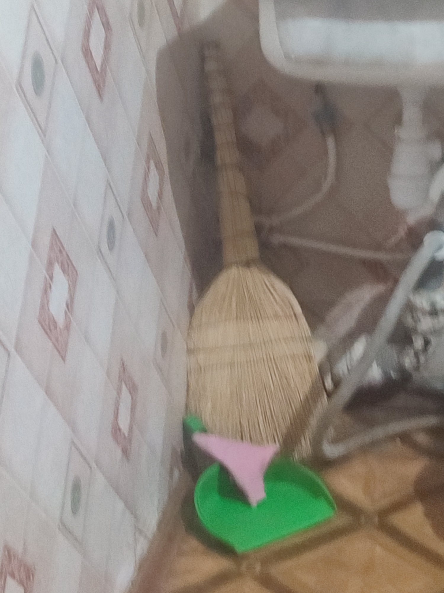 The image shows a broom and dustpan placed against a wall with a diamond-patterned tile. The broom has a straw-like texture and a wooden handle. The dustpan is green with a pink lip. The wall tiles are predominantly white with a light pink diamond pattern and some dark circular accents within the diamonds. The floor appears to be tiled in a wood-grain pattern. In the background, partially visible, are a white appliance, possibly a washing machine or a toilet tank, and some tubing. The scene is likely in a domestic setting, such as a bathroom or kitchen, in Chernivtsi, Ukraine.