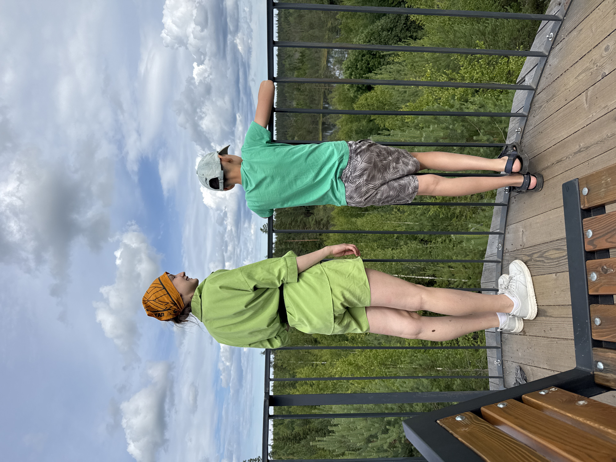 Two people, a young female and a young male, stand on a wooden platform, looking out at a forest landscape under a cloudy sky. The female, on the left, faces away from the camera and wears a green linen outfit consisting of a collared shirt and shorts, an orange bandana on her head, and white sneakers. The male, on the right, also faces away from the camera, wearing a green t-shirt, patterned shorts, a light-colored baseball cap, and dark sandals. They are positioned behind a black metal railing. In the foreground, a wooden bench with black metal supports is visible. The background is a dense forest with various shades of green trees and a body of water in the distance. The sky is filled with large, white cumulus clouds. The location is identified as the city of Peski, Russia.