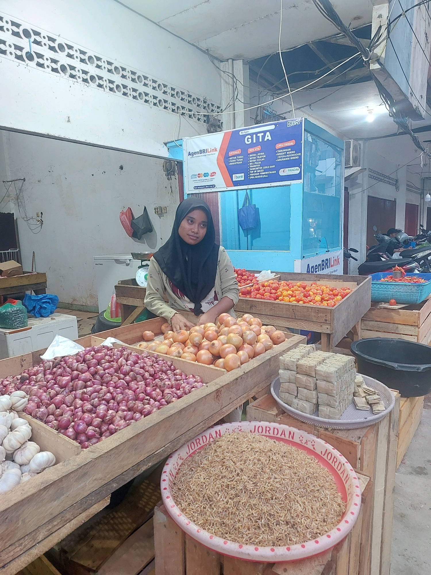 An outdoor market stall in Ambon, Indonesia, features a young woman wearing a black hijab and a patterned shirt, tending to a display of produce. In the foreground, crates overflow with shallots and garlic. Behind these, a crate of tomatoes and a blue basket of chili peppers are visible. To the right, a black plastic tub sits next to a display of bundled items wrapped in palm leaves. In the background, a blue counter with promotional signage for "GITA" and "AgenBRIlink" is set up, suggesting a financial services point. Various motorbikes are parked behind the stall, and the overall environment appears to be an open-air market with a concrete floor and a corrugated metal roof.