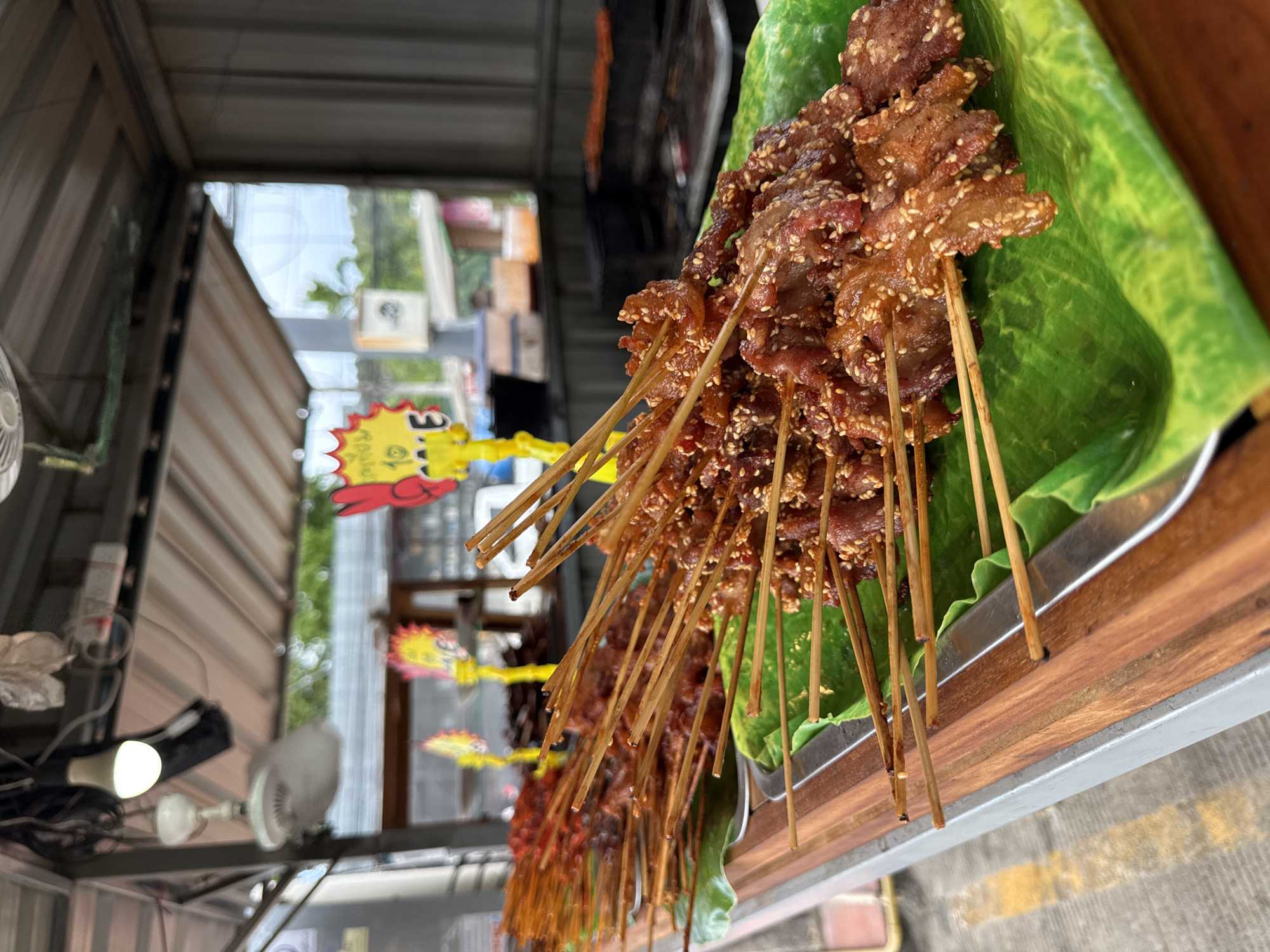 In Baan Bo Phut, Thailand, a close-up shot focuses on several skewers of grilled meat displayed on a table. The meat, coated in sesame seeds, is arranged on bright green plastic leaves, which are themselves placed in silver trays. These trays rest on a wooden surface, part of a market stall. In the background, a grill with more skewers of food is visible, along with yellow and red signs with text. The overall scene depicts a food vendor's setup, likely in an outdoor market setting. The lighting suggests it is daytime.