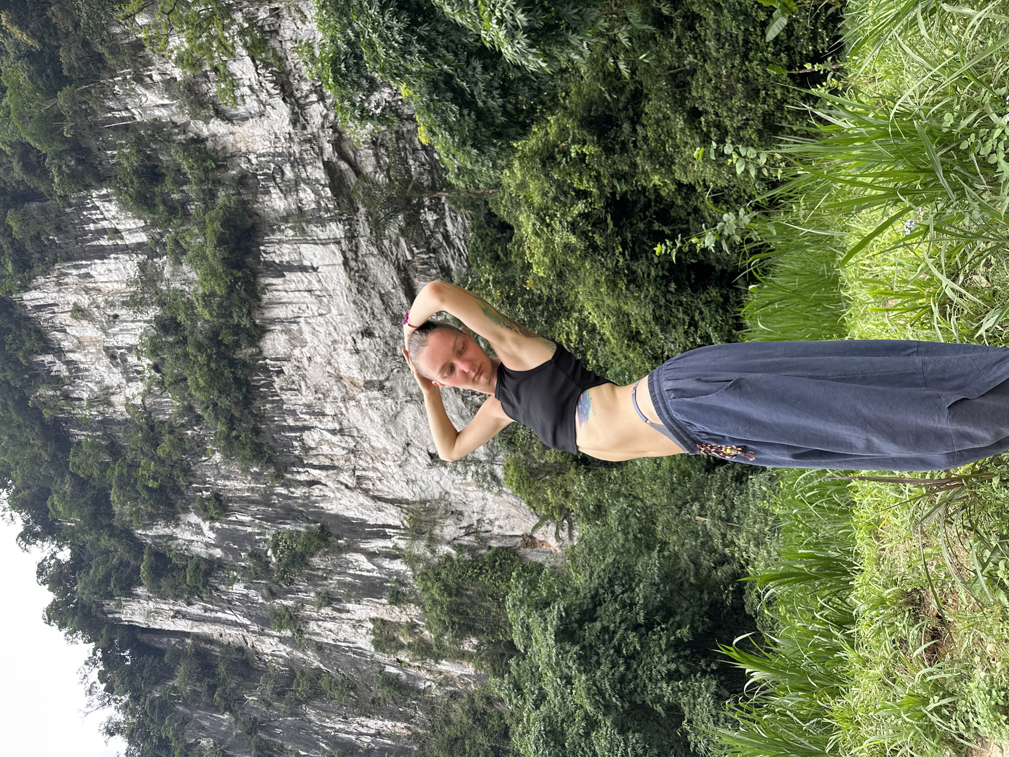 A young woman with fair skin and blonde hair pulled back stands in a natural environment in Pù Luông Commune, Vietnam. She is wearing a black crop top and dark blue baggy pants. Her arms are raised above her head, and her body is angled to the side, revealing a tattoo on her hip. The background features a towering, sheer rock face covered in lush green vegetation, with dense trees and tall grasses in the foreground. The sky is overcast.