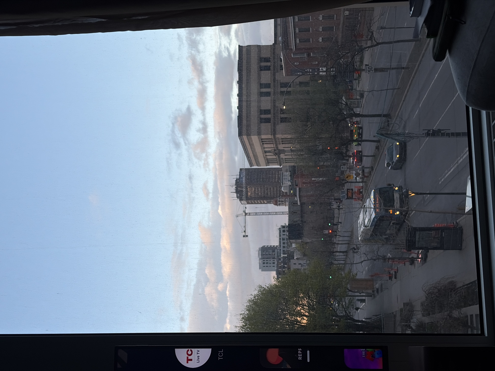 A slightly blurry, slightly tilted photograph looking out of a window at a city street scene. The sky is partly cloudy, and buildings of various sizes and architectural styles are visible along the street. Construction is evident with a crane near a high-rise building. Trees are present along the street. Traffic is moving along the streets including a bus. There is a TV visible at the bottom of the window. While difficult to confirm definitively, the architecture and general layout resemble a street in Montreal, Canada.