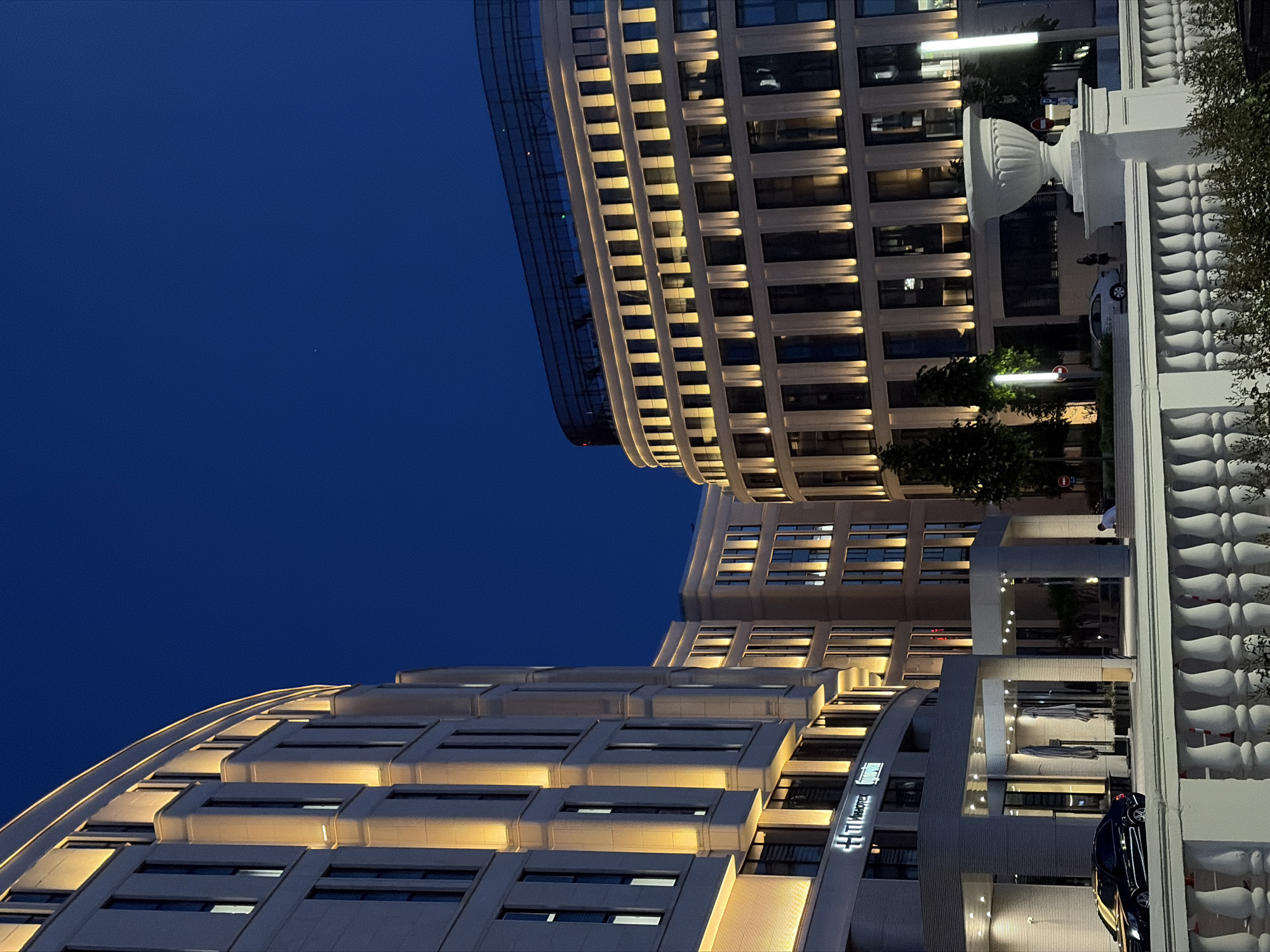 The photograph captures an evening scene in Minsk, Belarus, showcasing modern architecture illuminated against a deep blue sky. In the foreground, a white balustrade runs horizontally across the frame, separating the viewer from a street-level view of a hotel entrance. A black car is parked to the left of the entrance. The hotel building on the left is a multi-story structure with a curved facade, featuring regularly spaced windows that are lit from within, casting a warm glow. Vertical lighting strips highlight the architectural details of the building.
To the right, another building with a similar modern design and a curved upper section is visible. This building also features numerous illuminated windows and vertical light accents. In the background, trees and streetlights are sparsely present. A large white ornamental urn stands to the right of the balustrade, adding a touch of classical design to the contemporary surroundings. A few individuals are visible in the distance, suggesting normal activity in the area. The overall impression is one of an urban environment at dusk, with buildings artfully lit to create an elegant ambiance.
