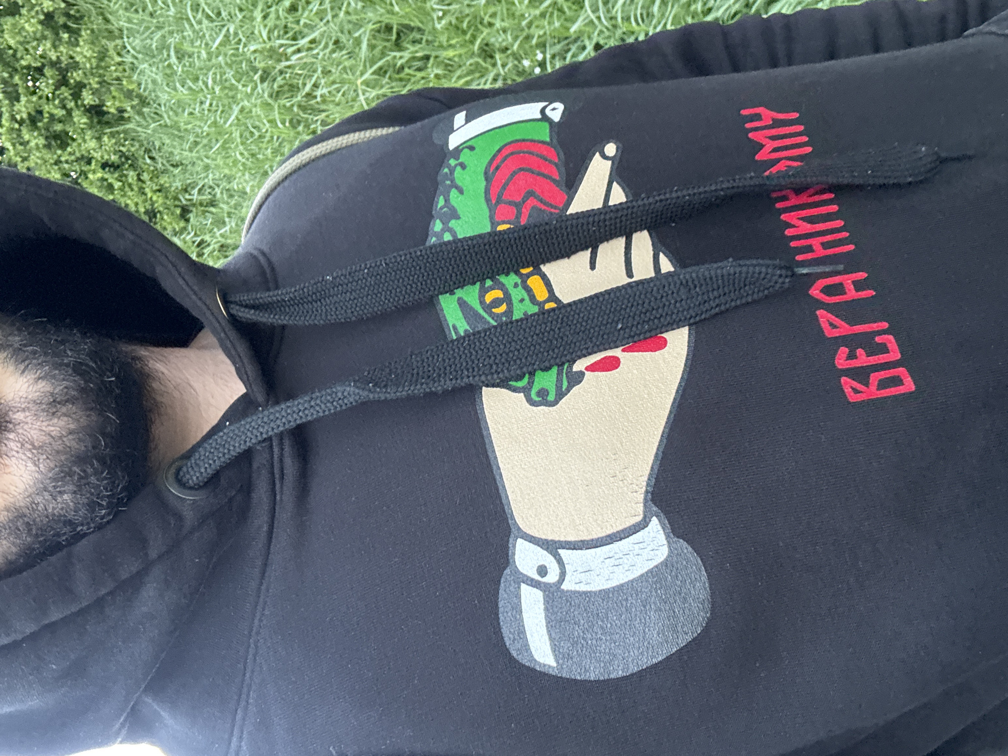 A man wearing a black hoodie lies on grass. The hoodie has a graphic of a hand holding a grenade. Vertical red text in Cyrillic script is next to the graphic. A black hood is visible on the left side of the frame. The man's face is obscured by the hood, revealing only his beard. The grass is green and appears to be well-maintained. The location cannot be definitively determined as being in Welshpool, United Kingdom based on the visual information provided.