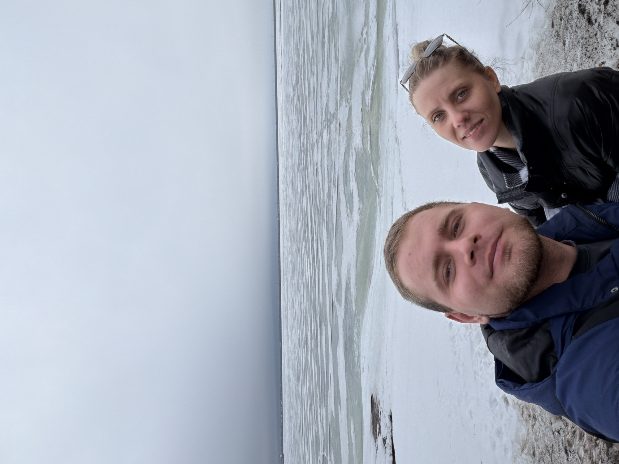 The image shows a selfie taken by two people, a man and a woman, possibly in the city of Chaikovsky, Russia, as the landscape resembles the Volga River or a large body of water typical of the region. The man is positioned in the foreground, wearing a dark blue jacket. The woman stands behind him, wearing a dark jacket with sunglasses perched on her head. They are both smiling. The background shows a grey sky and a snow-covered landscape, possibly a frozen body of water, or the bank of the Kama River.