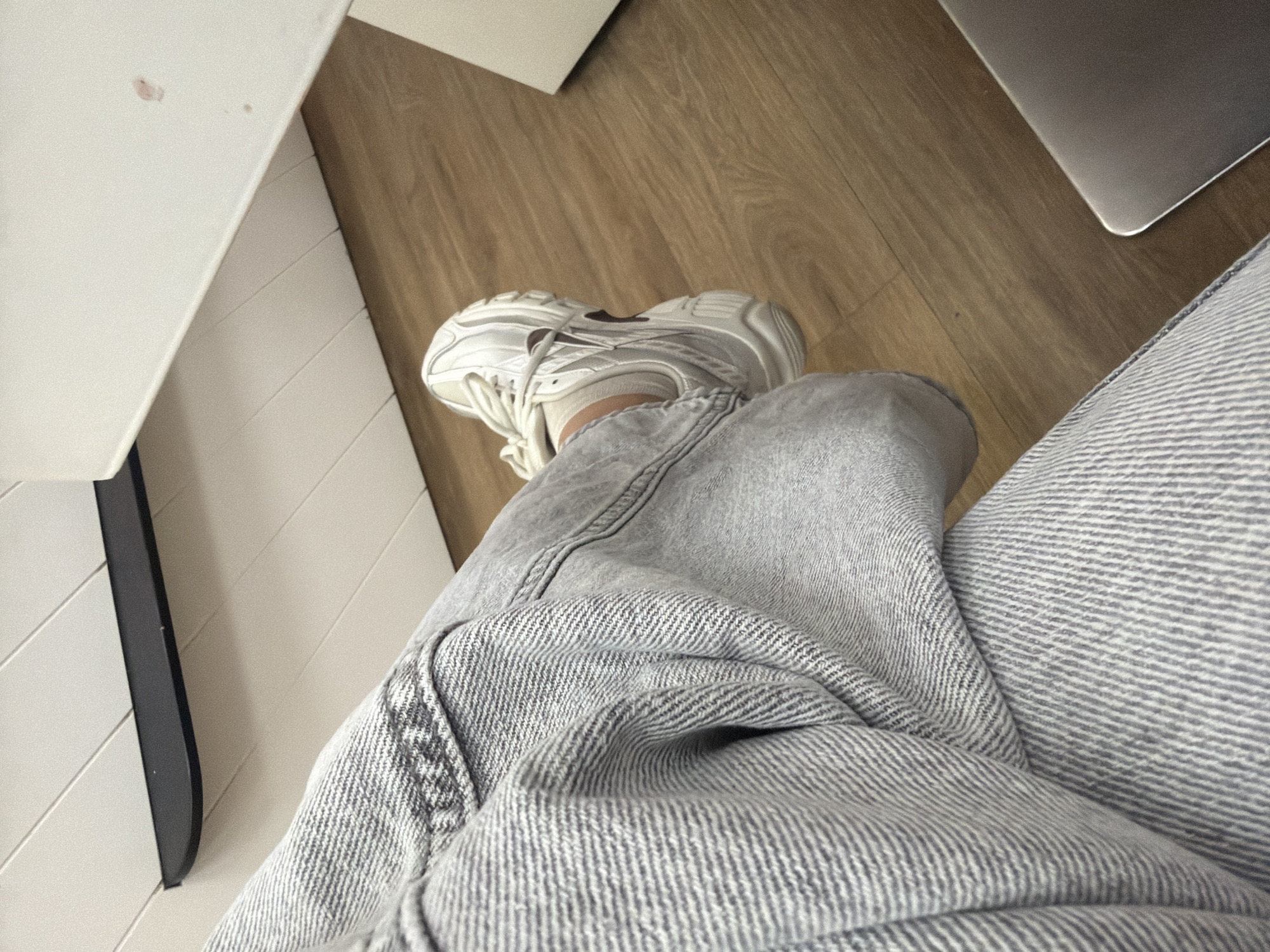 The image presents a first-person, close-up view of a person seated, presumably indoors. The subject is wearing light gray jeans and a white sneaker. The sneaker sits on a wooden floor, next to a white table. A laptop is visible in the top right corner. There is no information to ascertain the location as Plovdiv, Bulgaria or any other city.